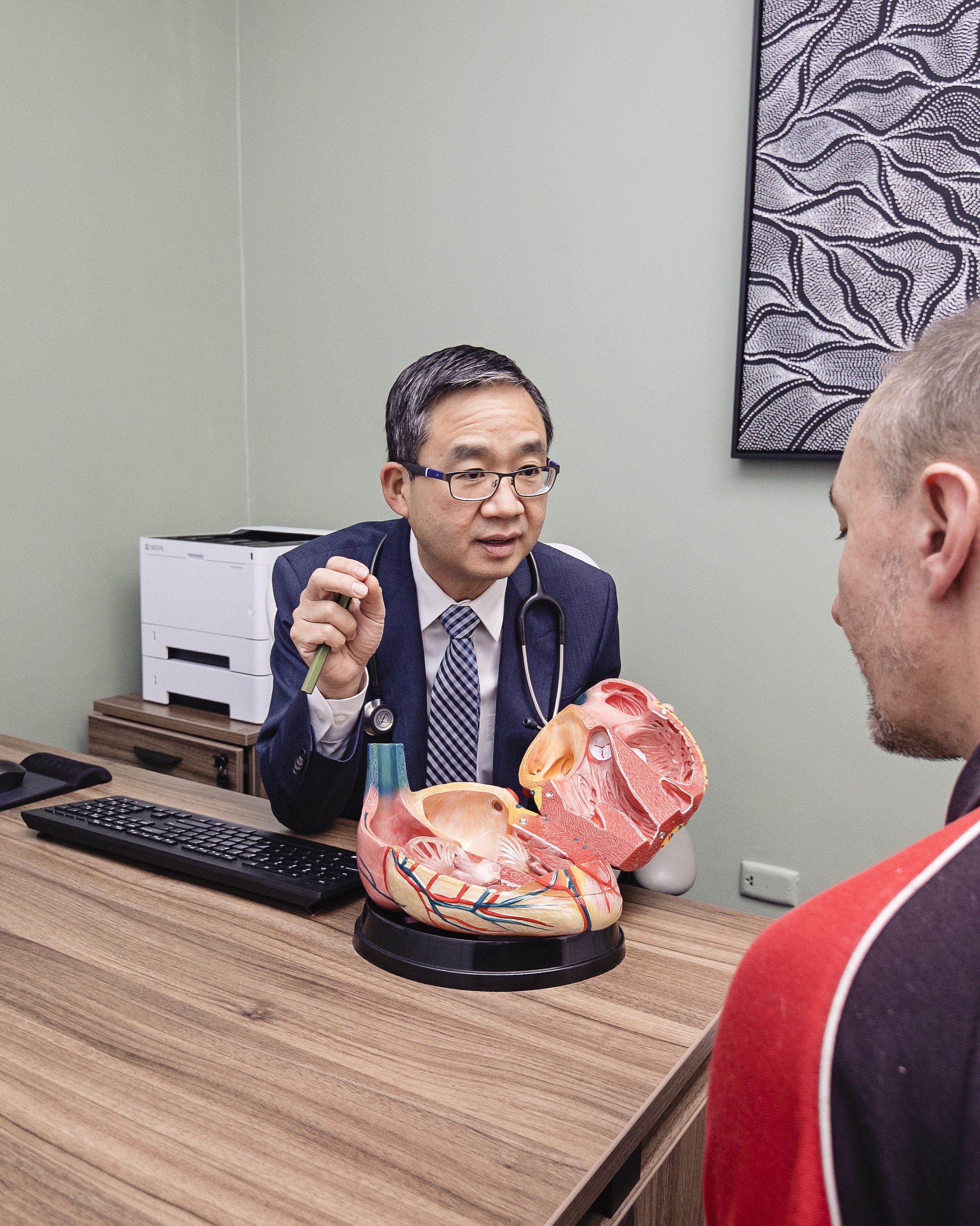 A doctor, wearing a suit and tie, sitting at a desk with a computer keyboard in front of him. The doctor is holding a pen and gesturing while talking to a patient, who is partially visible and wearing a red sweater. On the desk, there is a detailed anatomical model of a human head showing the internal structures, including muscles, nerves, and blood vessels. The background features a piece of abstract art on the wall, a white medical printer, and a wood-textured office surface.