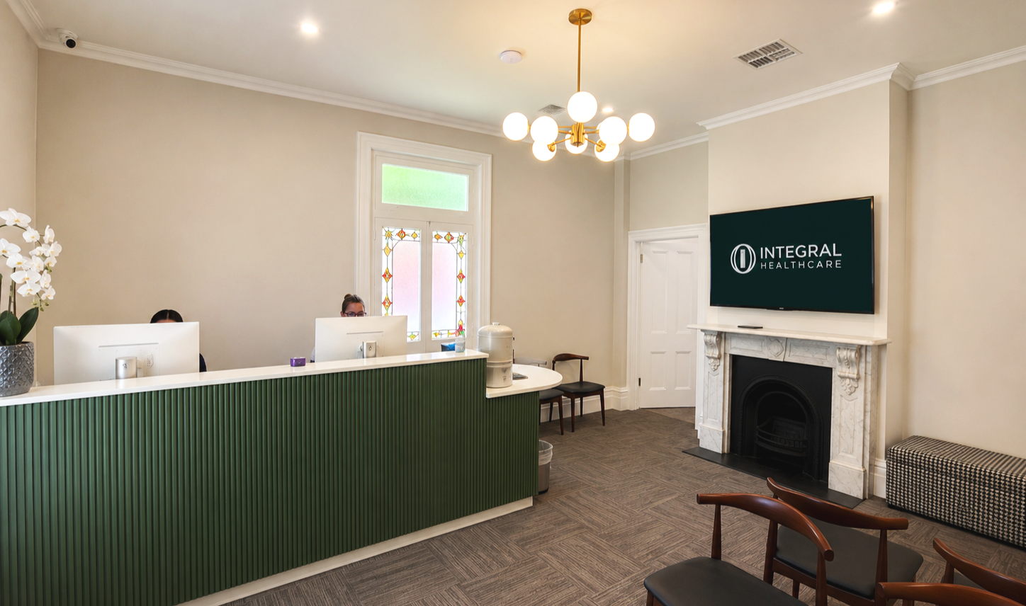 Reception area with a green front desk, two people working at computers, a fireplace below a large screen displaying 'Integral Healthcare,' a chandelier, and a window with stained glass details.