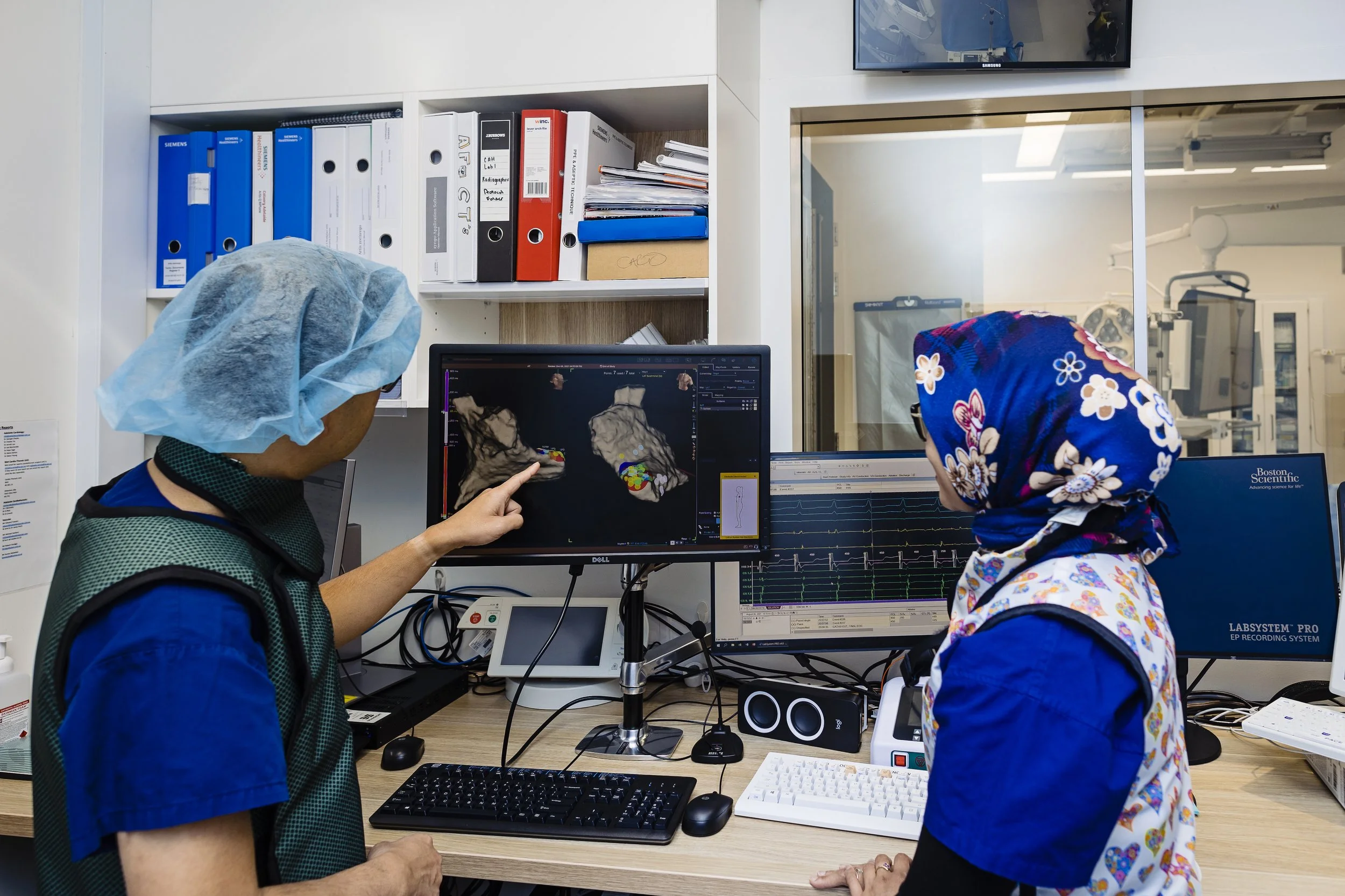 Two healthcare professionals wearing surgical caps and scrubs are examining medical scans on computer monitors in a clinical setting. One person is pointing at the screen, while the other observes.
