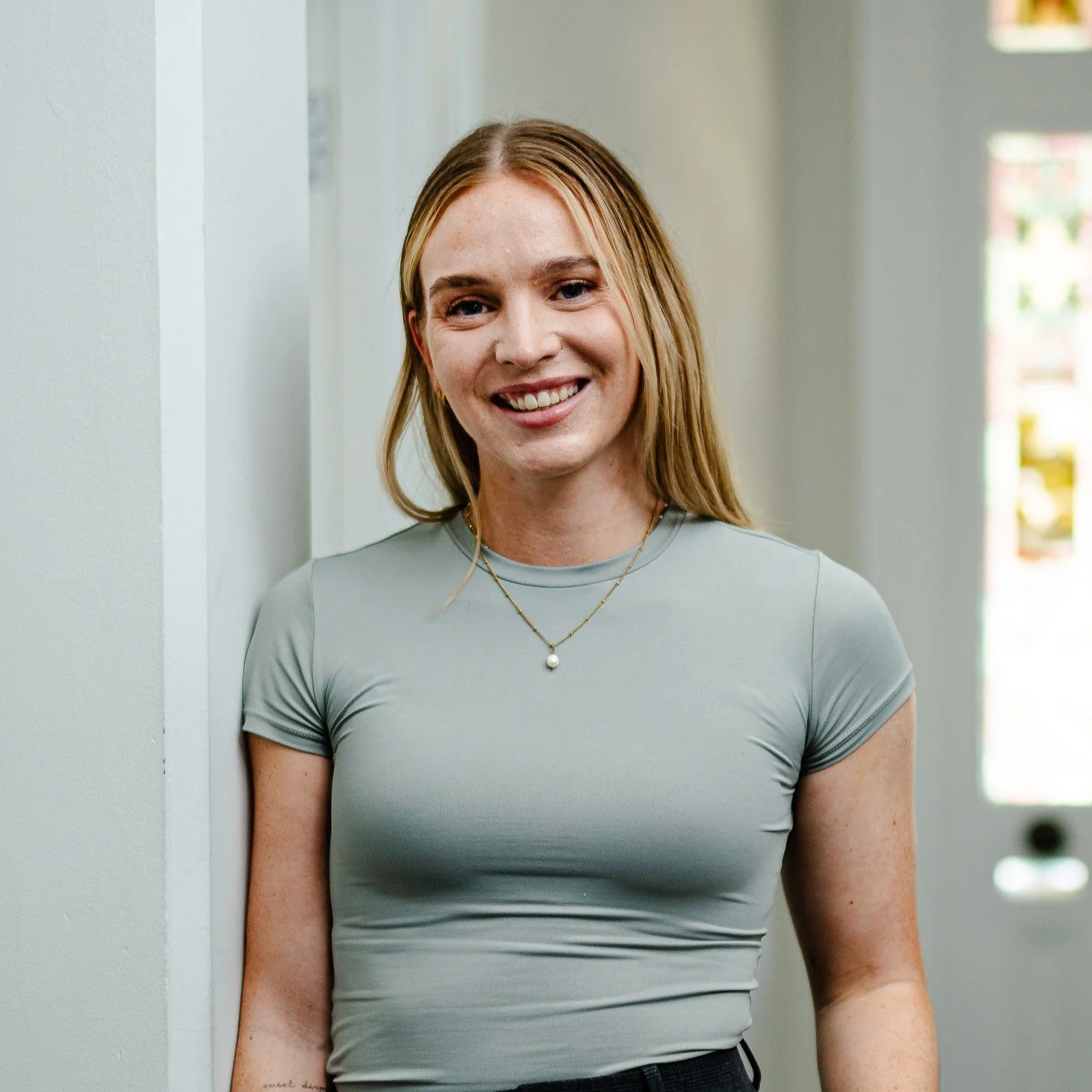 A smiling woman with long blonde hair, wearing a light gray fitted t-shirt and a delicate necklace, standing against a white wall near a window.