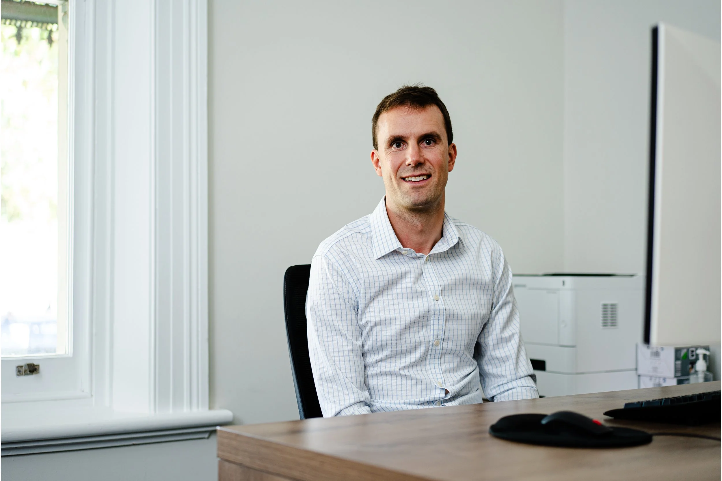 A man with short dark hair wearing a light-colored checkered shirt sitting at a desk in an office with a window and white cabinets in the background.