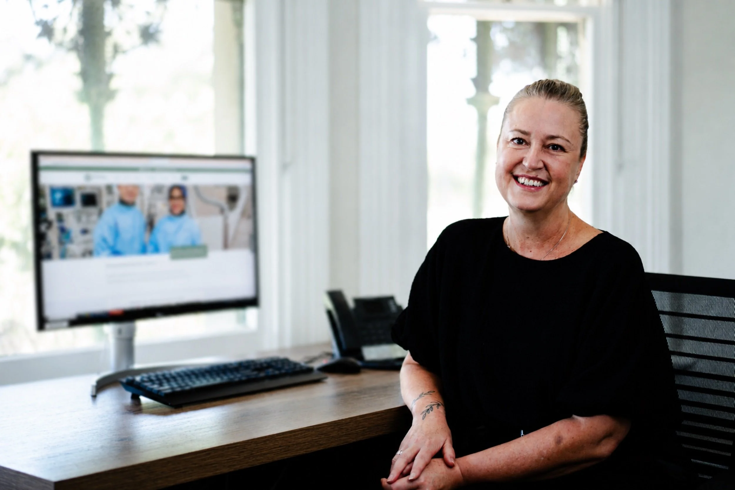 A woman with blonde hair and tattoos on her arm smiling in an office. In the background, a computer monitor displays two people in medical gowns, and there is a large window letting in natural light.