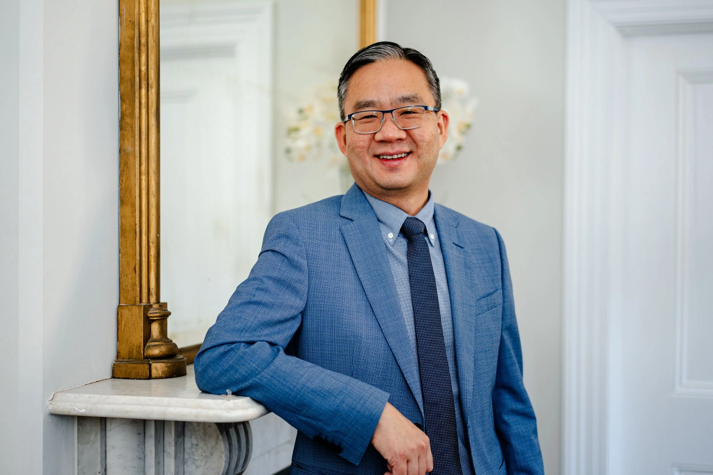 A man in a blue suit and glasses smiling while standing next to a mirror and leaning on a white marble fireplace mantel in a well-lit room.