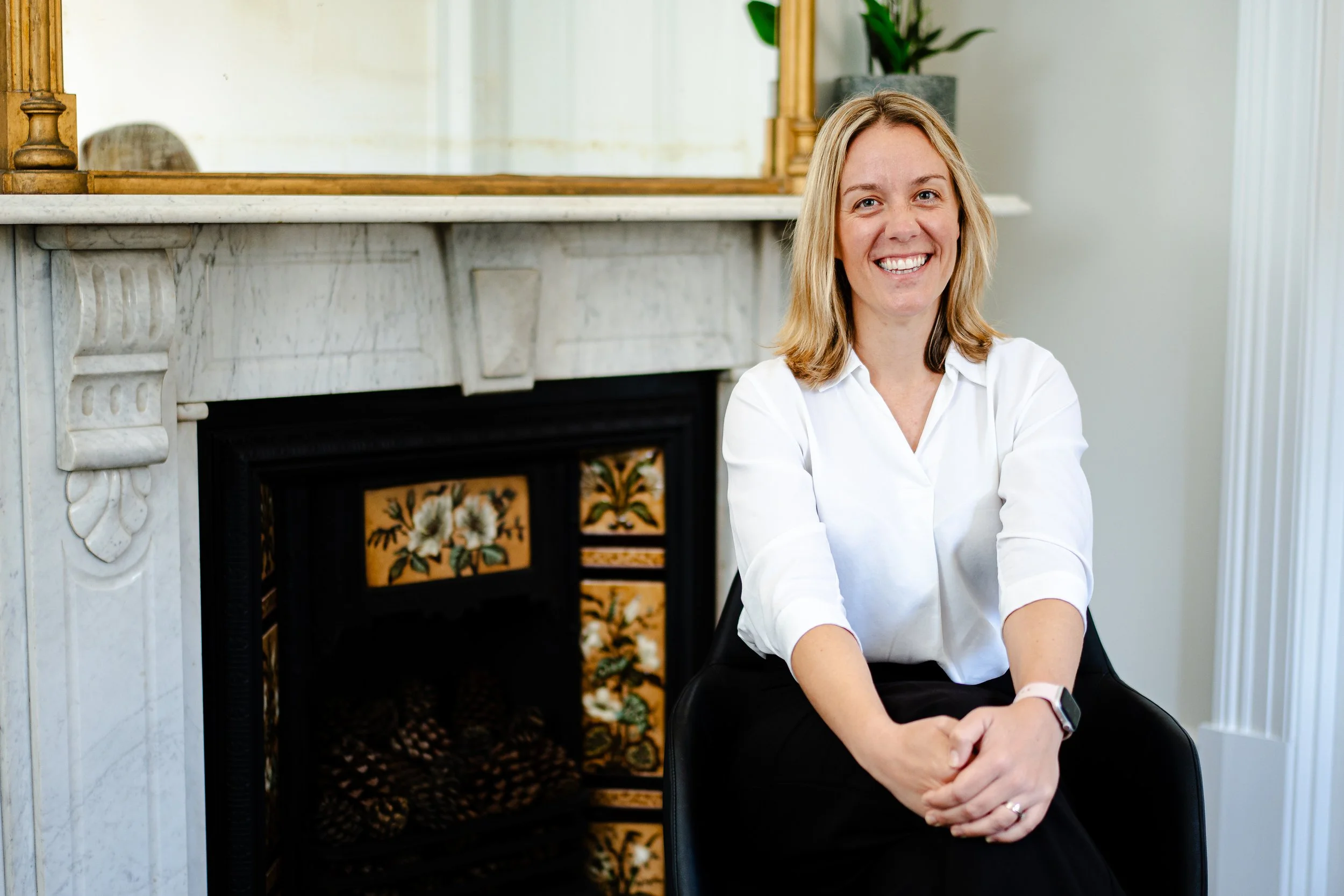 A smiling woman with blonde hair and a white shirt sitting in front of a marble fireplace with ornate details, and a large mirror hanging above it, in a room with white walls and a plant.