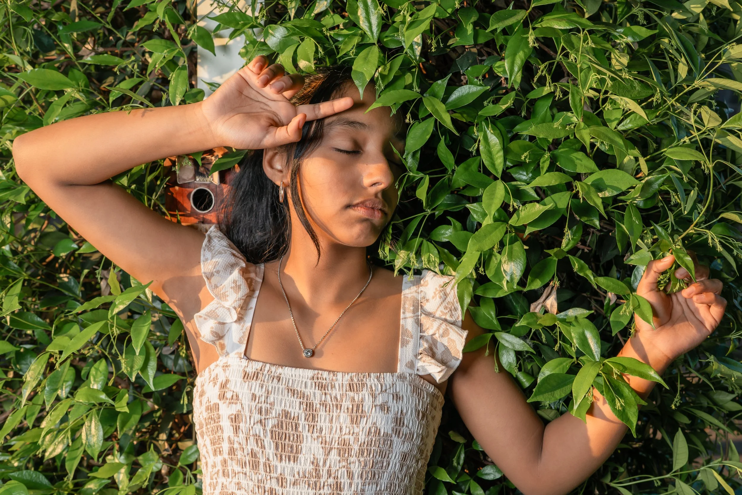 A young woman with dark hair and earrings lying among green leaves, with her eyes closed and one hand near her face.
