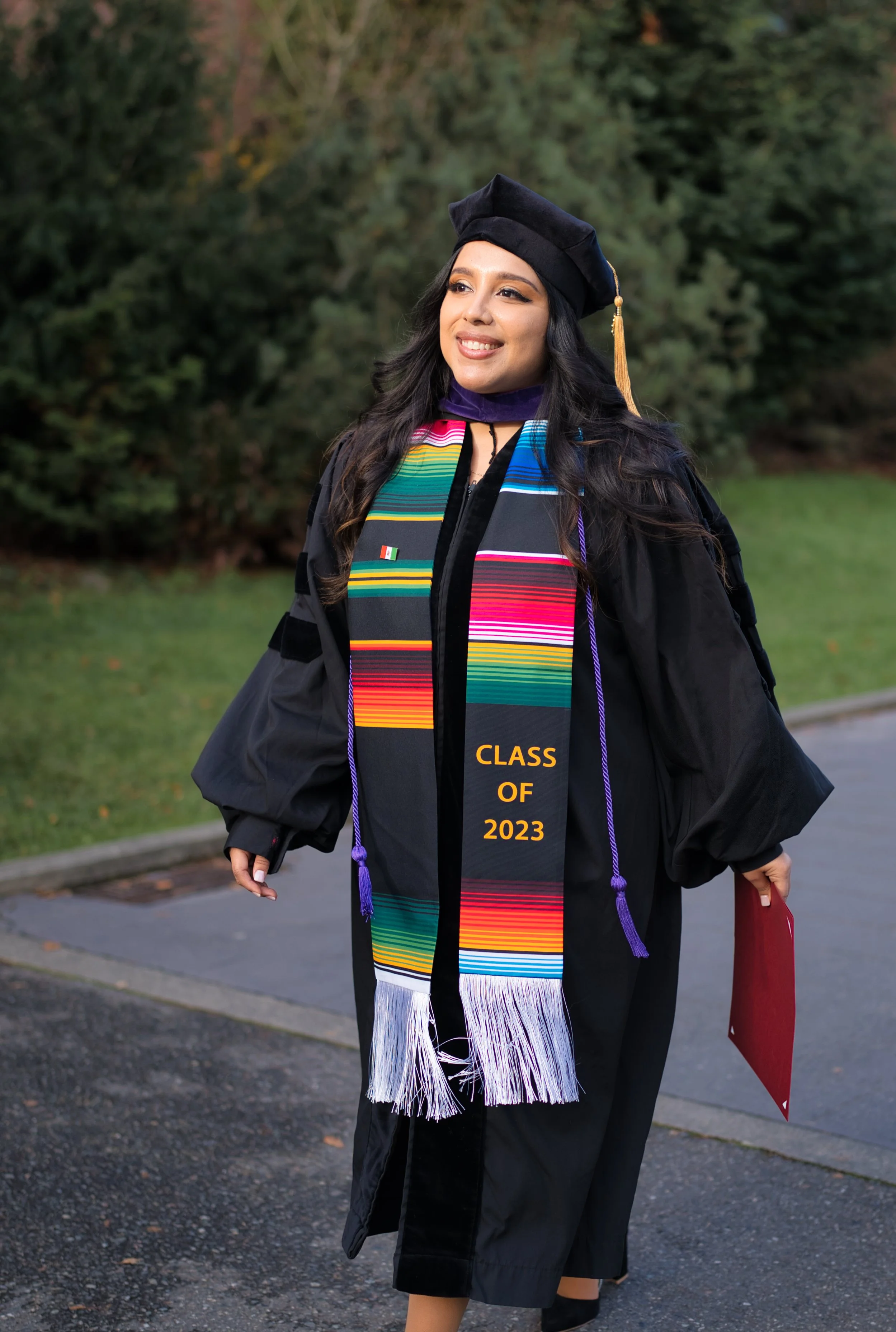 A young woman dressed in graduation cap and gown, wearing colorful stole and sash that reads 'Class of 2023', standing outdoors with a background of trees and grass during daytime.