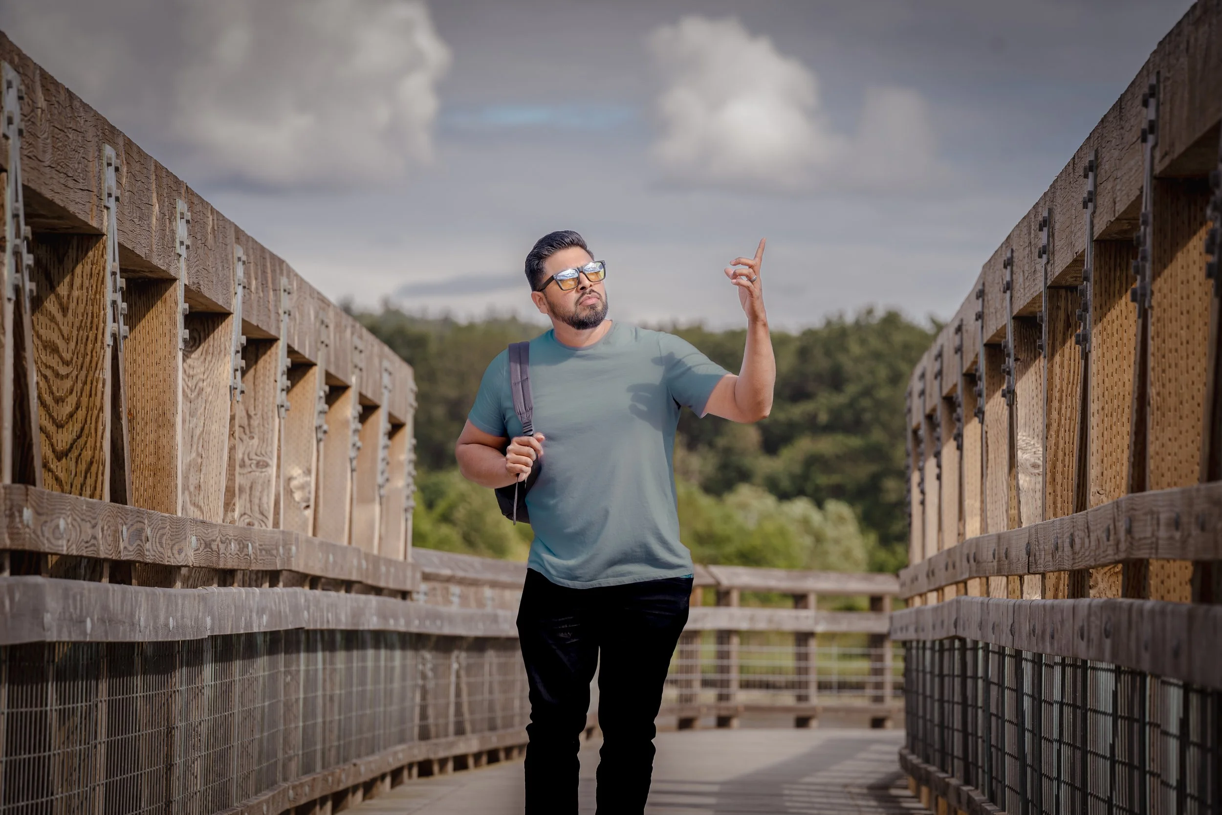 A man with sunglasses, a beard, a gray t-shirt, and a backpack walking on a wooden bridge with railings, pointing upward with one finger under a cloudy sky.