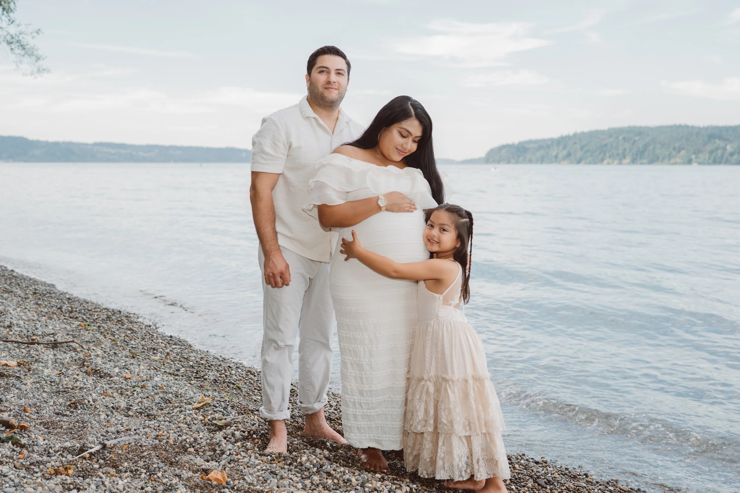 A family of three standing on a pebble beach near a lake with hills in the background. The woman, who is pregnant, is smiling and looking down at her belly, while the man stands behind her, smiling at the camera, and a young girl hugs the woman around her waist.