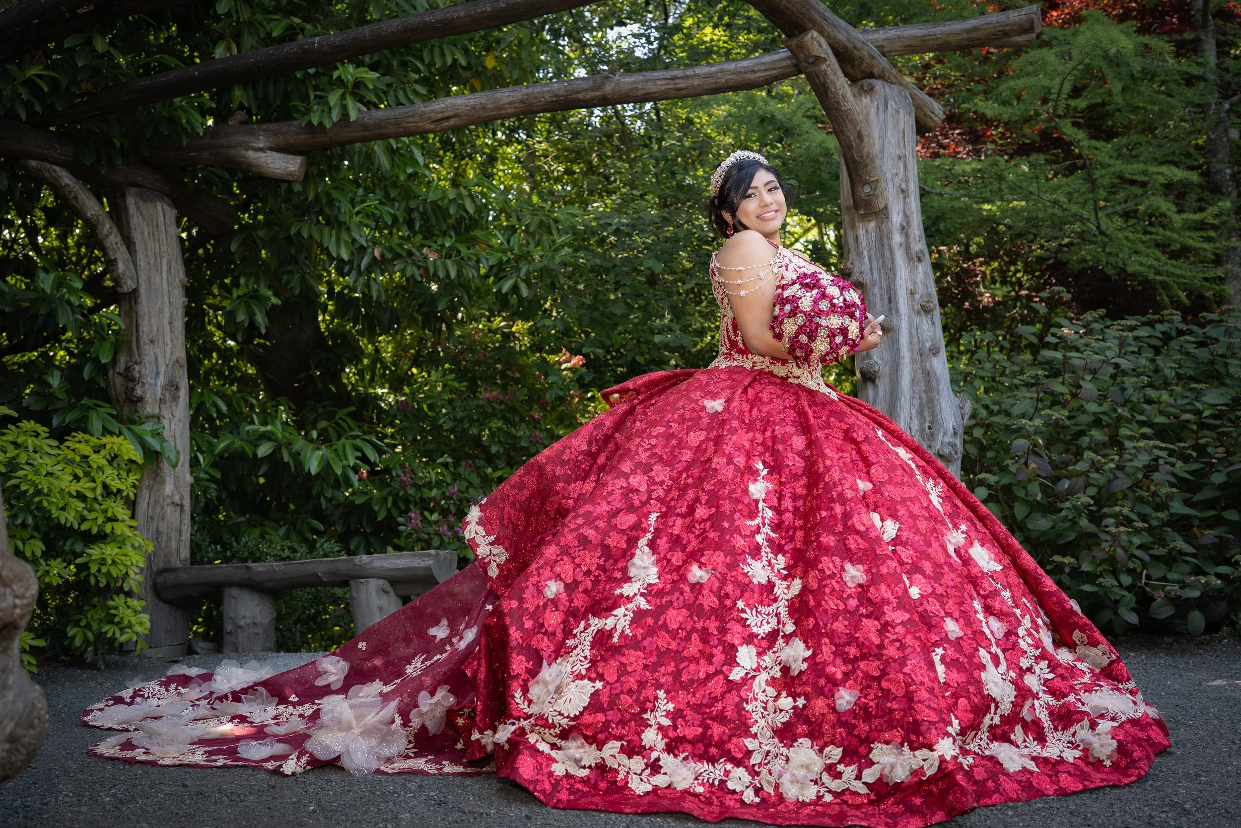 A woman in a red and white floral bridal gown and tiara standing outdoors near trees.