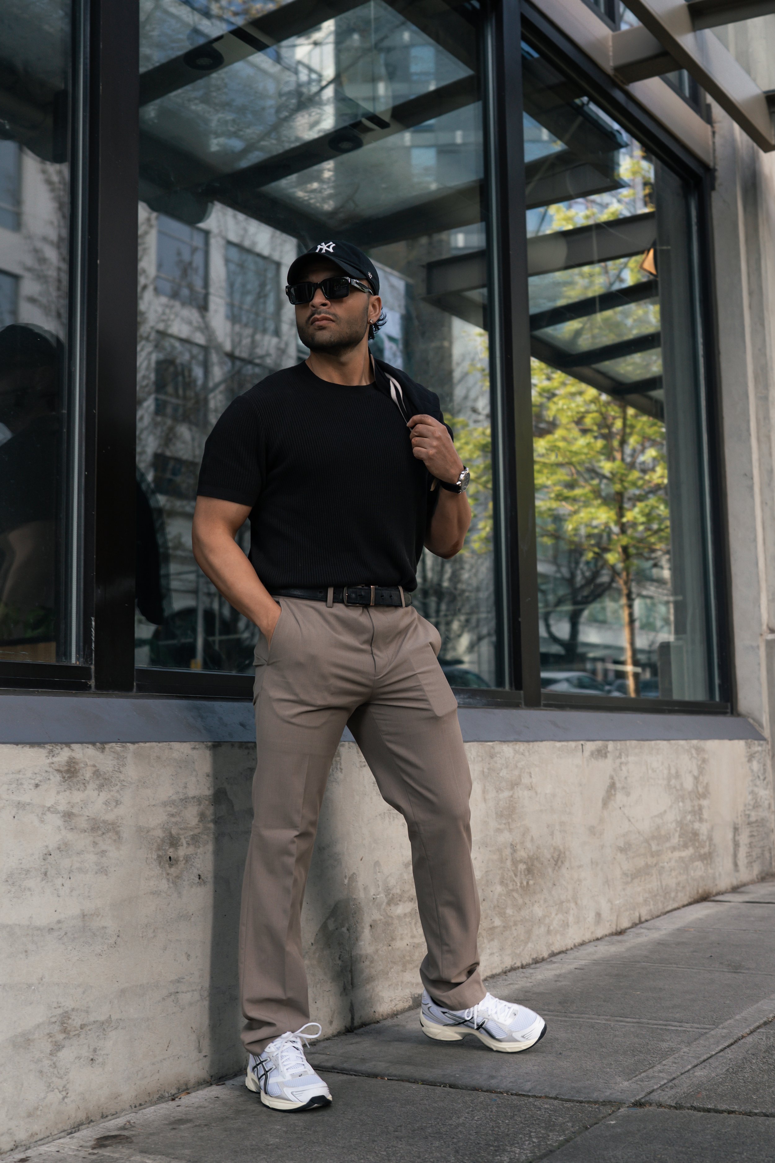 A man wearing a black T-shirt, beige pants, white sneakers, a black cap, and sunglasses is standing outside near a building with large glass windows. He has a backpack over one shoulder and has one hand in his pocket. Trees and a building are reflected in the glass.
