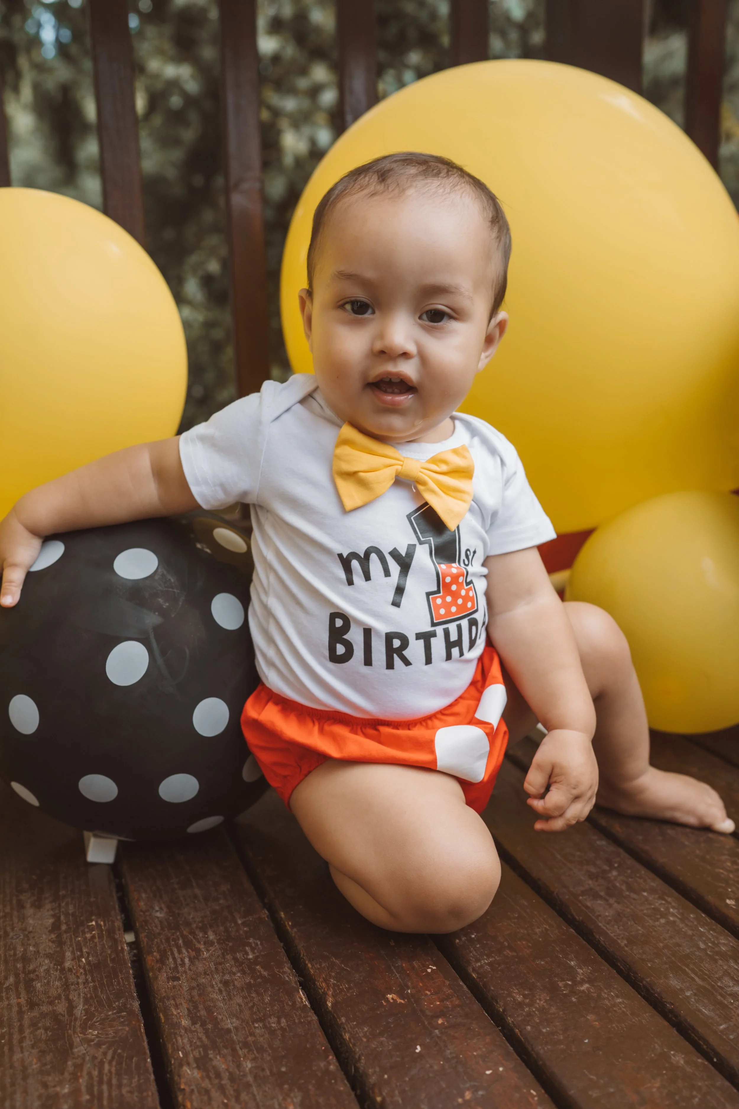 A young child celebrating a birthday, sitting outdoors on a wooden deck surrounded by large yellow, black, and white polka dot balloons. The child is wearing a white t-shirt with a birthday message and a yellow bow tie, along with red shorts with white polka dots.