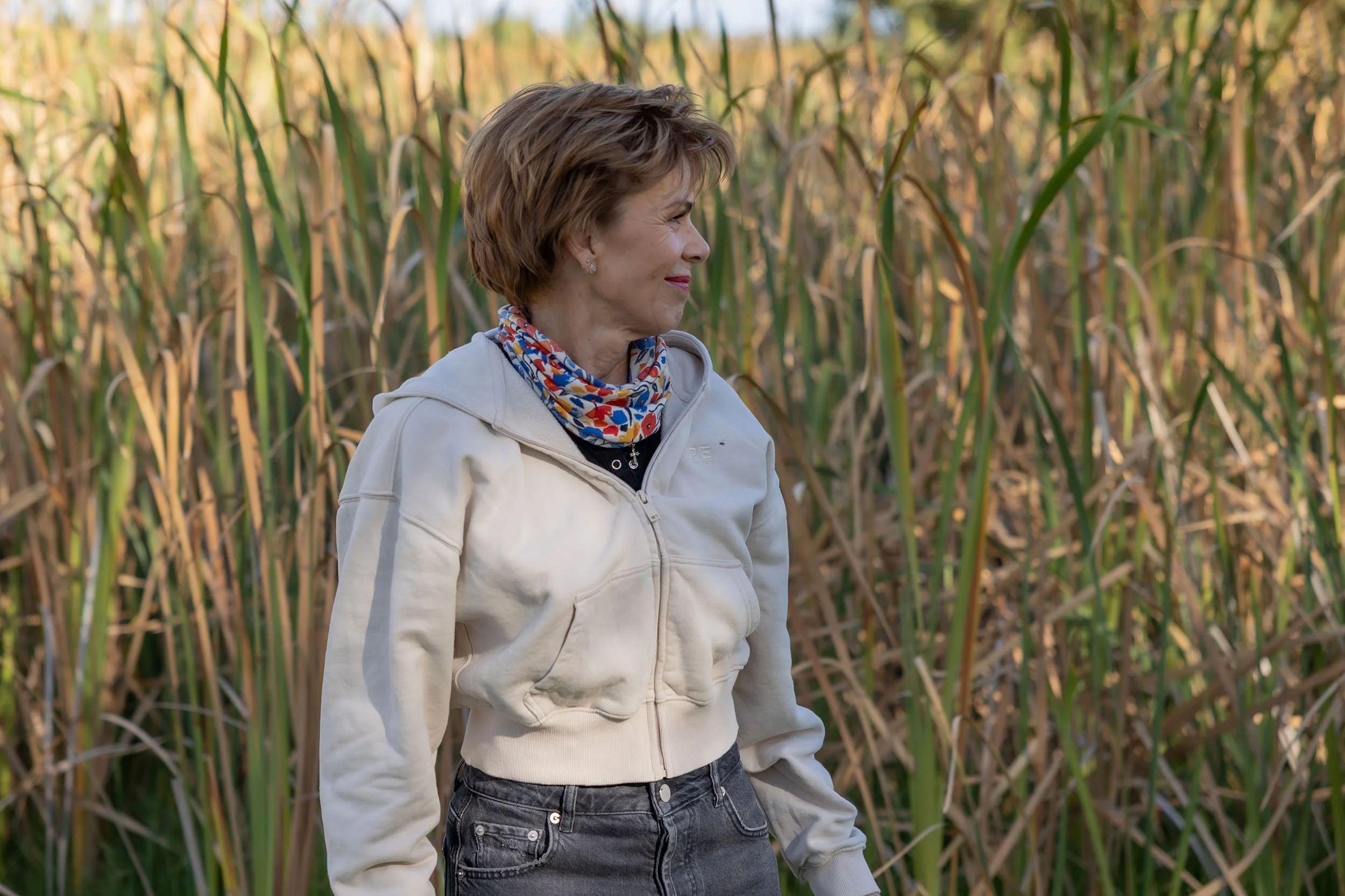 A woman standing in a cornfield during daylight, wearing a light-colored jacket and a colorful neckerchief, looking to her right.