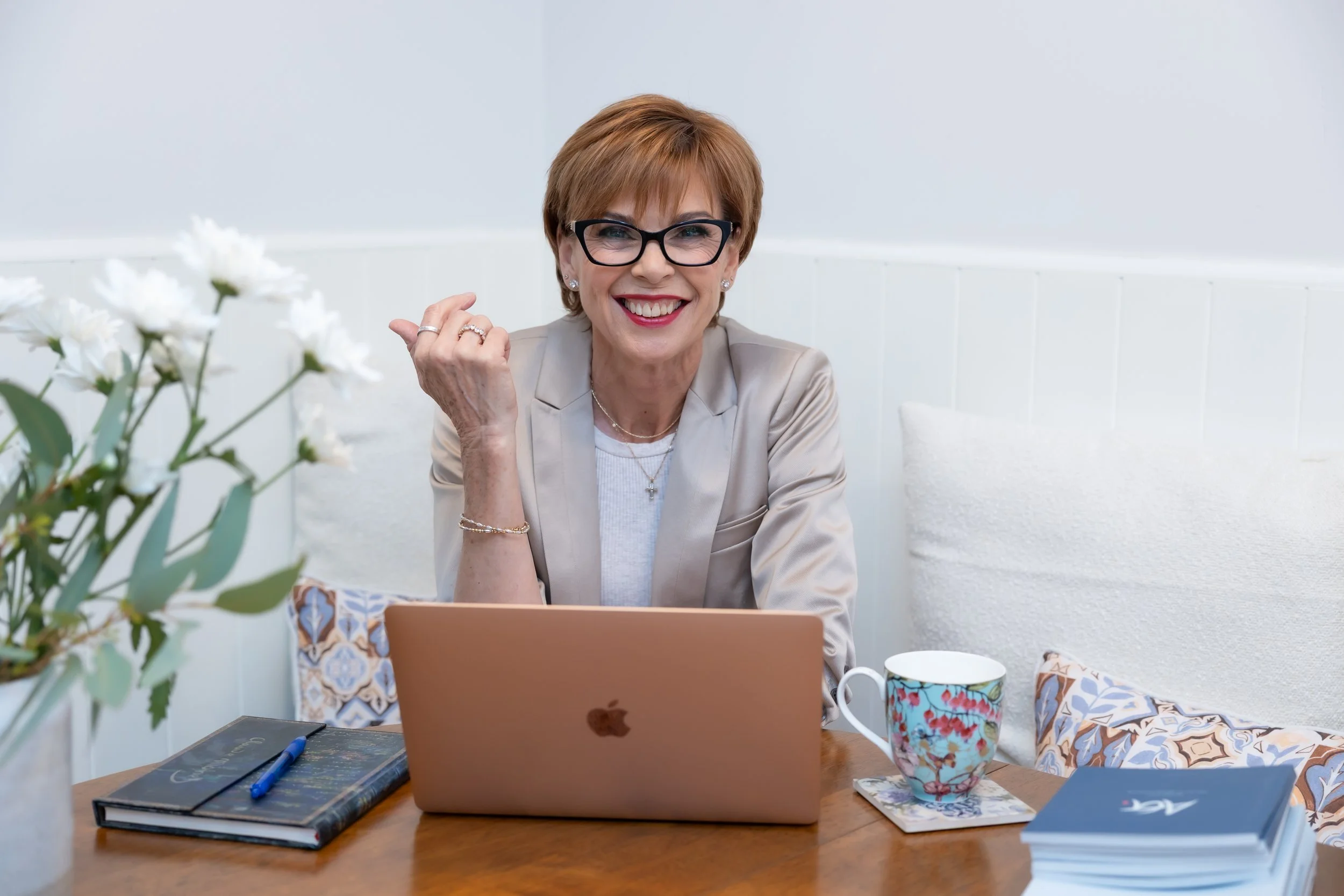 A smiling woman with short red hair, glasses, and jewelry sits at a wooden table with an open laptop, surrounded by notebooks, mugs, and decorative items in a bright, white room.