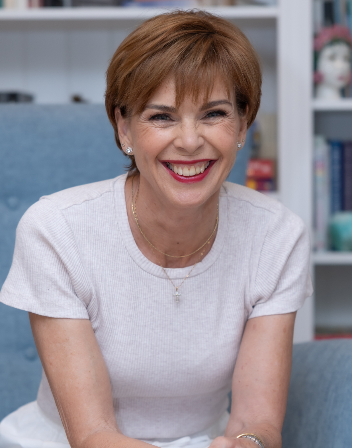 A woman with short red hair, smiling broadly, sitting indoors with a background of bookshelves.