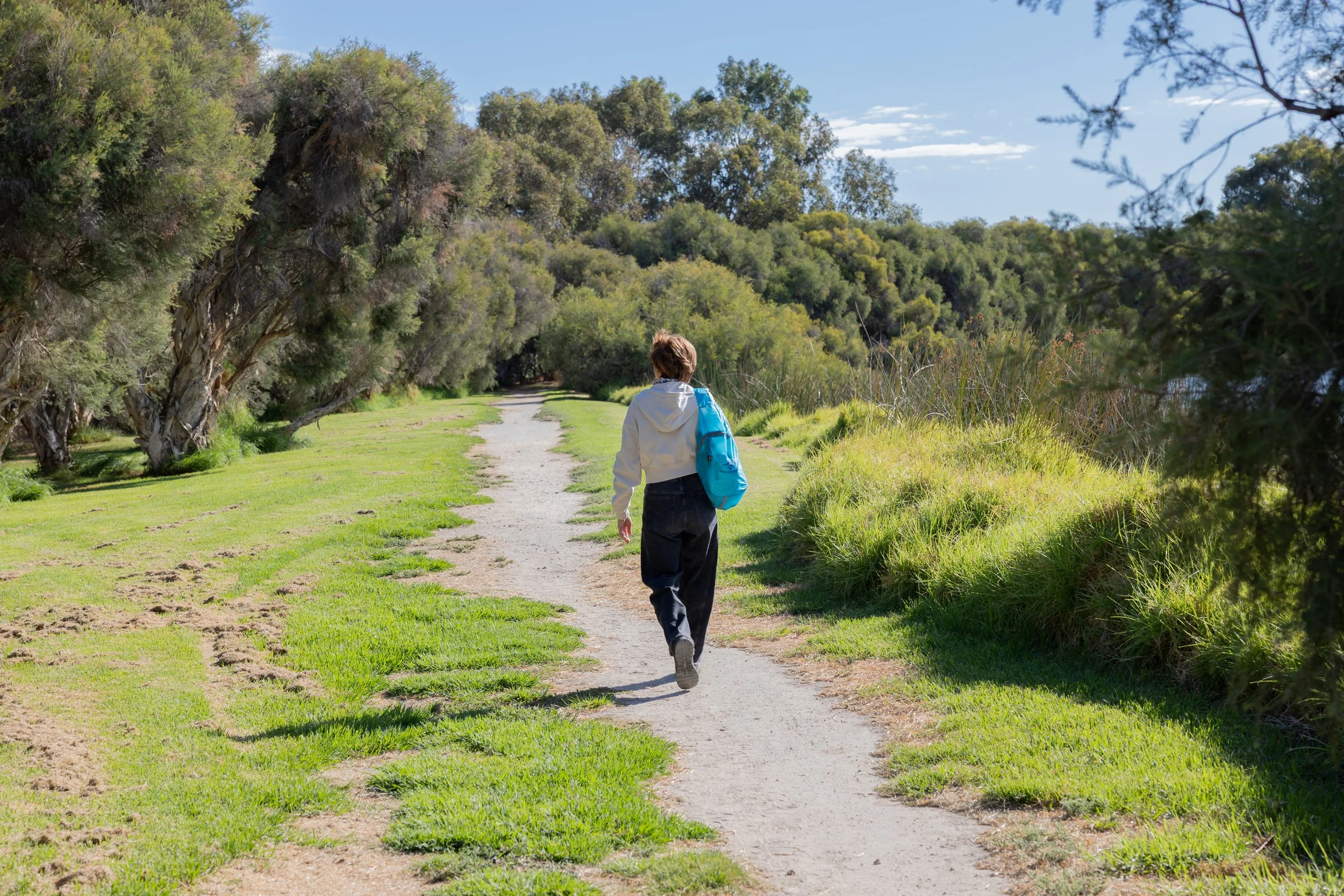 A person walking on a dirt trail surrounded by green trees and grass on a sunny day.