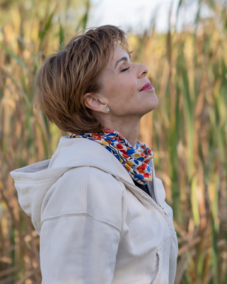 Woman with short hair and a colorful scarf standing outdoors in a field of tall grass, with her eyes closed and a peaceful expression.