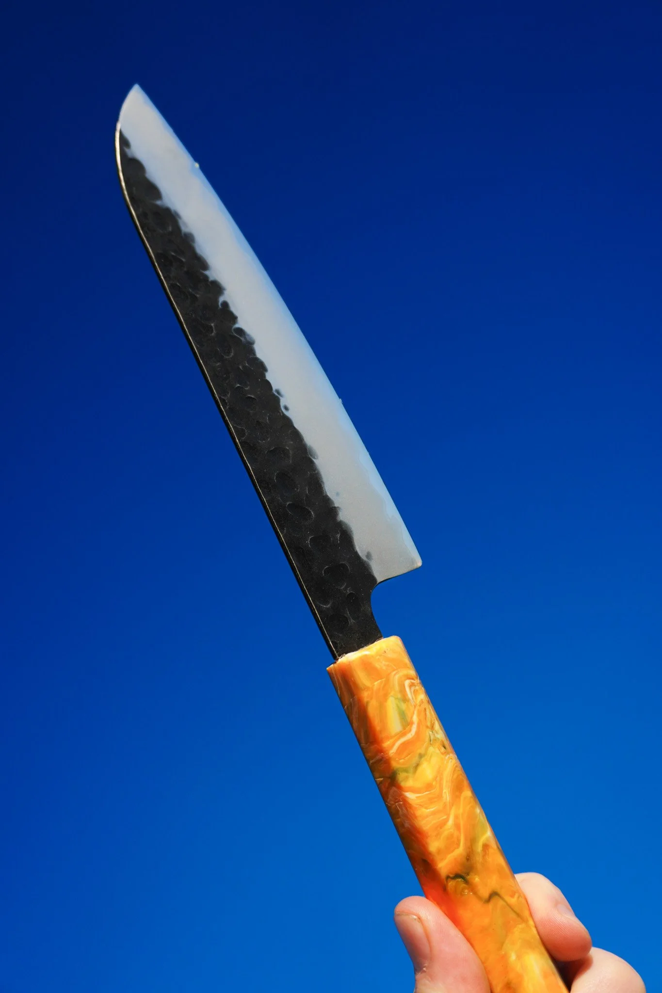 A hand holding a chef 's knife with a black and white patterned blade and an orange and yellow marbled handle against a bright blue sky.
