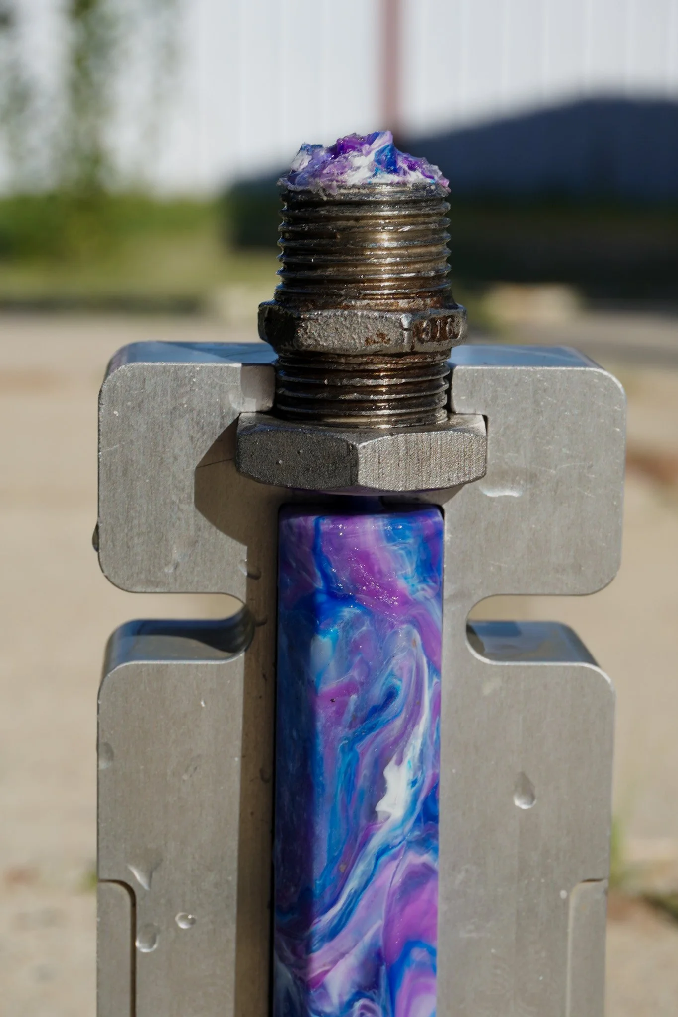 Close-up of a metallic bolt with purple, blue, and white paint swirling around it, mounted on a piece of machinery outdoors.