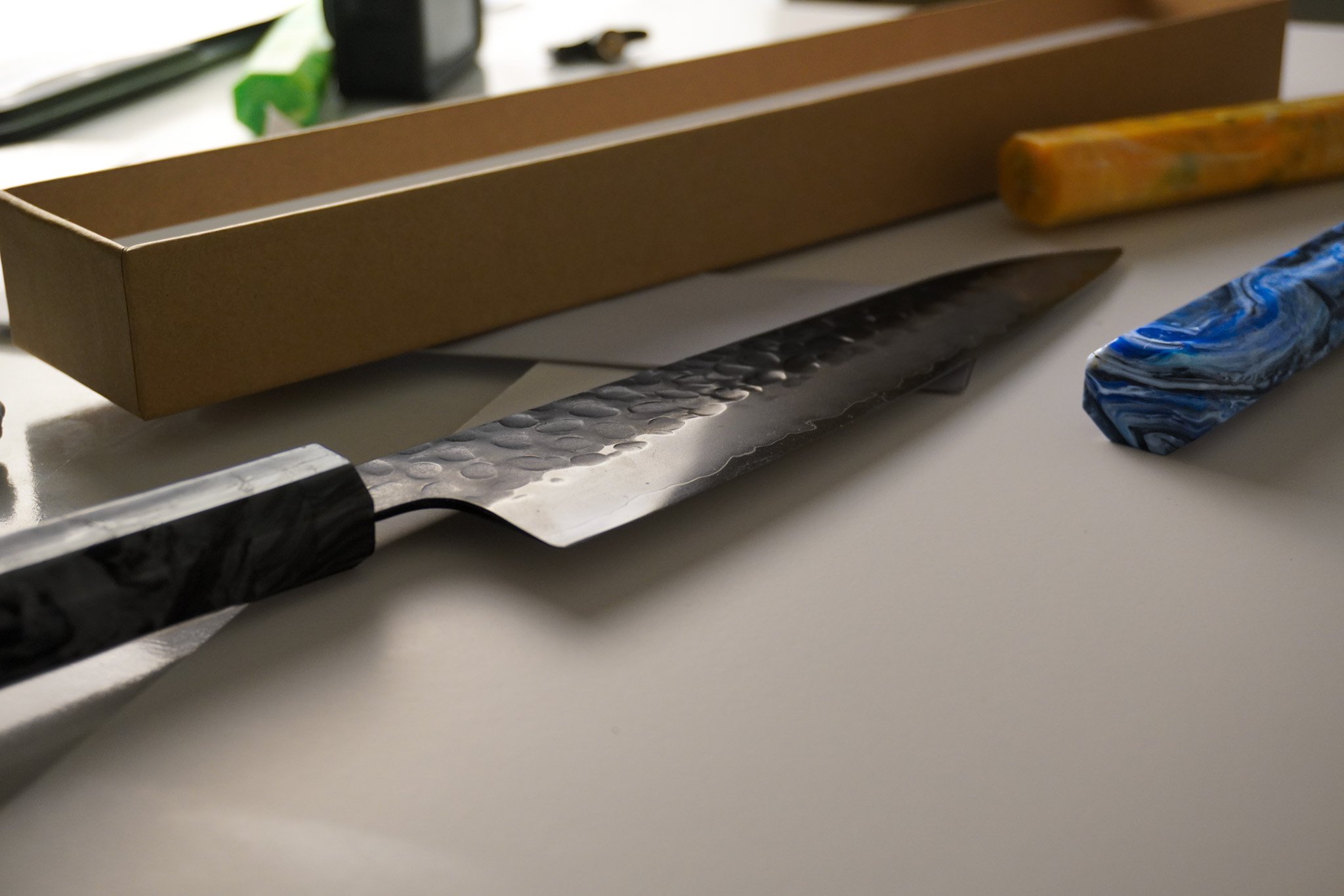 A cutting tool with a black handle, a serrated blade, a black and white marbled handle, a blue marbled object, a brown cardboard box, and various small tools and objects on a white surface.