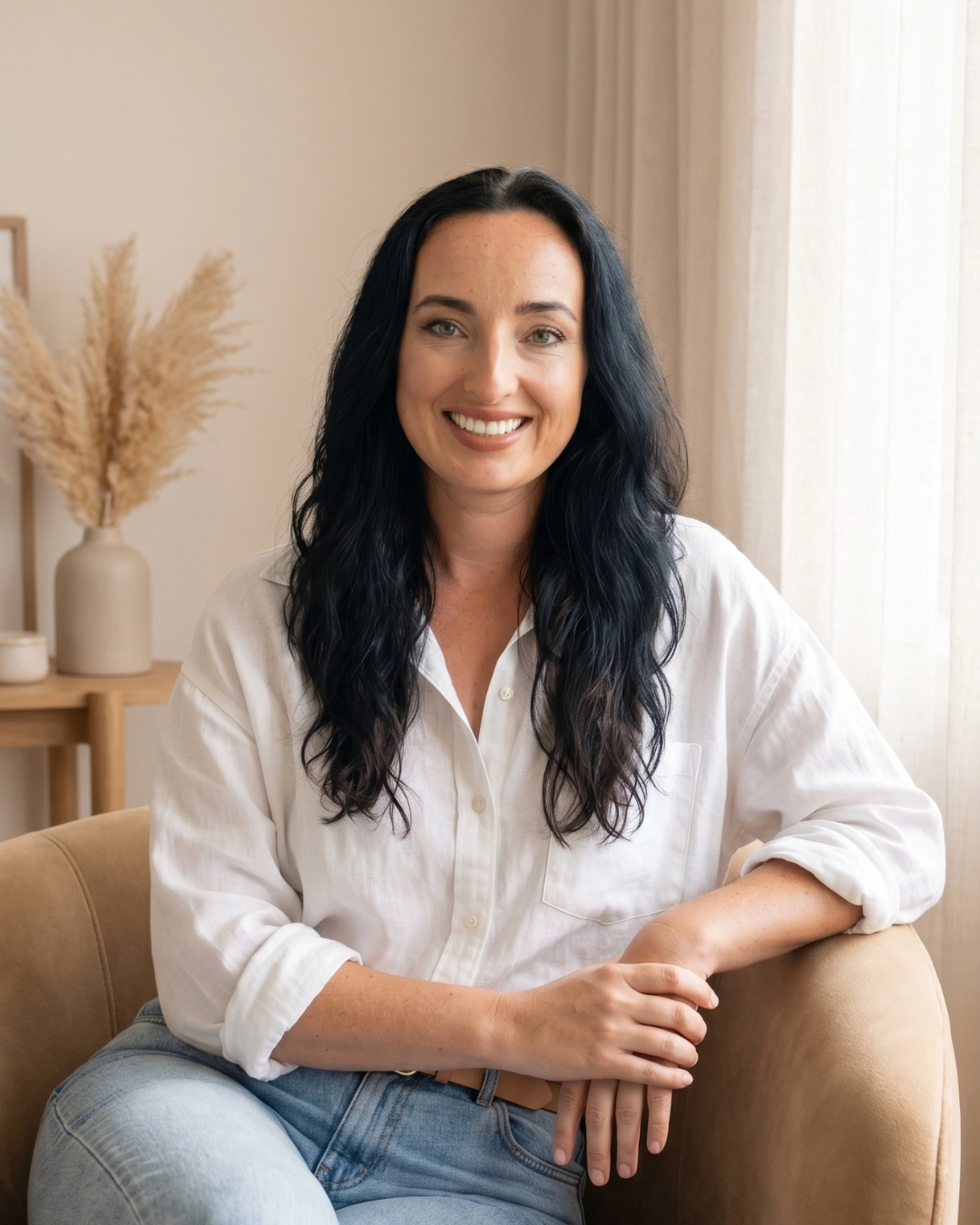 A woman with long, wavy black hair smiling and sitting on a beige couch in a well-lit room with curtains and decorative vases with pampas grass in the background.