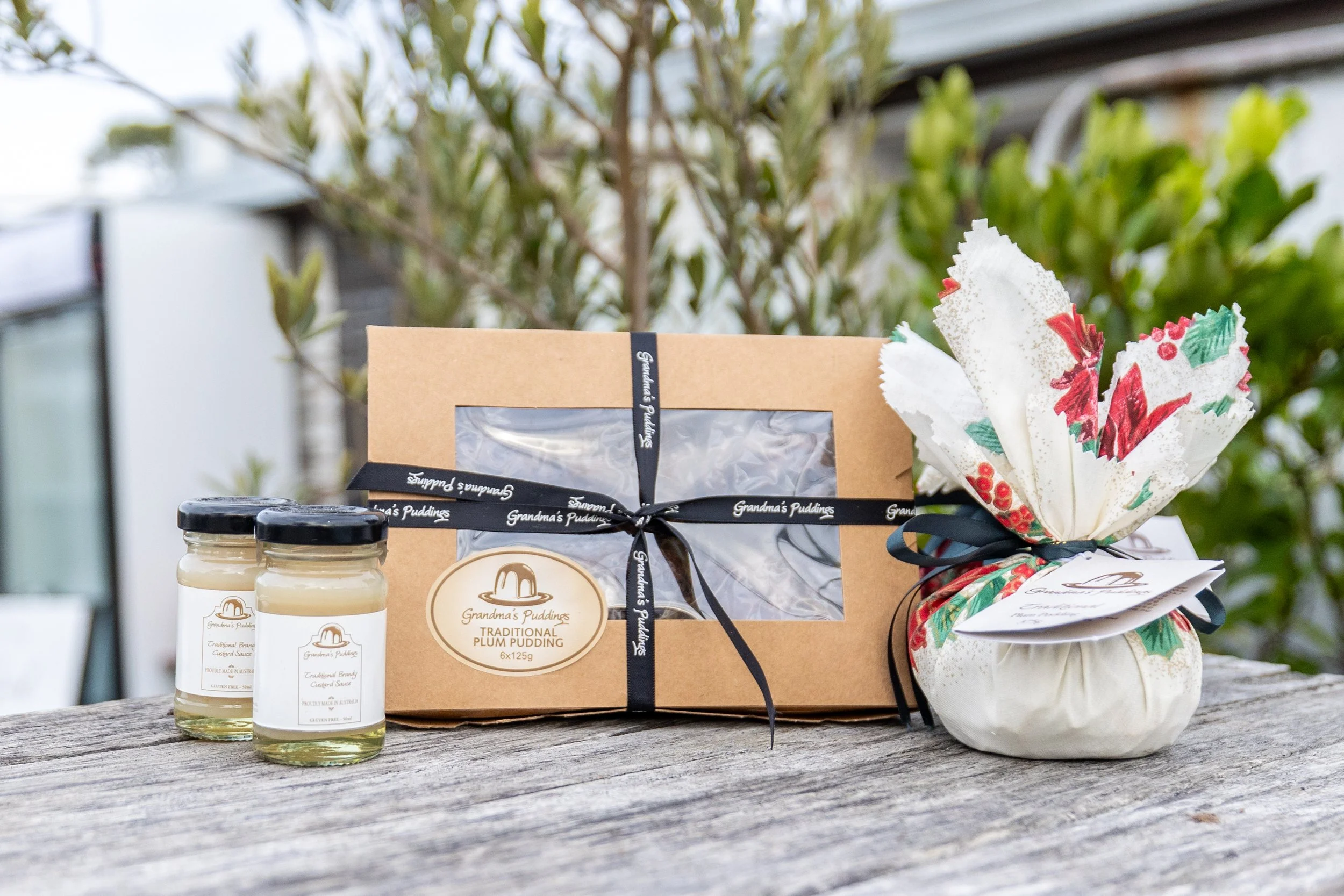 A gift box labeled Grandma's Puddings Traditional Plum Pudding, along with two jars of custard sauce, a wrapped gift or decoration with Christmas-themed paper, and two jars of custard sauce on a rustic wooden surface with a blurred outdoor background.