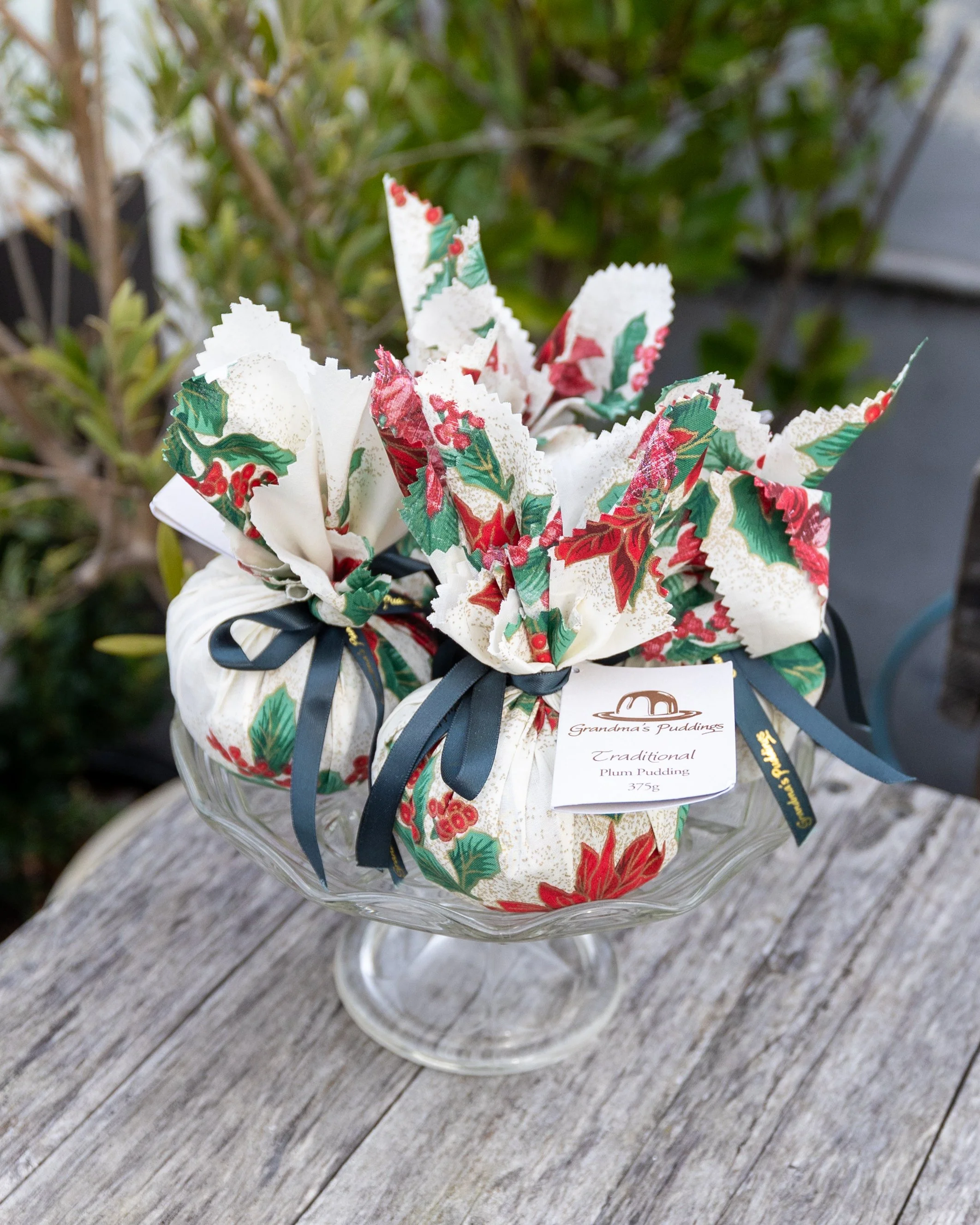 A decorative glass bowl filled with paper napkins wrapped in festive holiday-themed paper with holly, berries, and Christmas flowers, tied with dark blue ribbons, and a tag that reads "Grandma's Puddings, Traditional Plum Pudding, 375g," placed on a weathered wooden surface with greenery in the background.