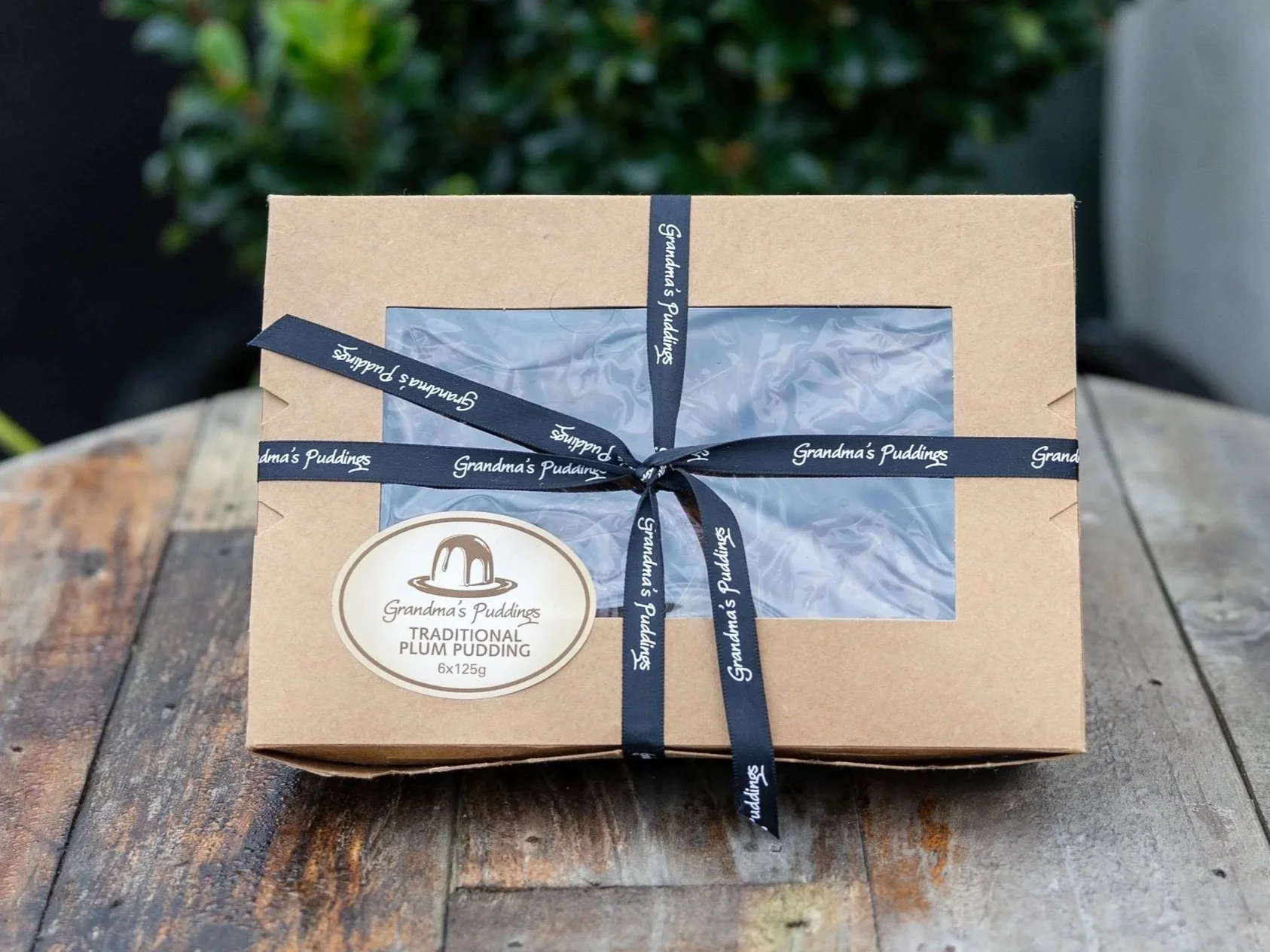 A rectangular box of Grandma's Puddings Traditional Plum Pudding with a transparent window on the top, tied with a dark ribbon that has 'Grandma's Puddings' written on it, placed on a wooden surface with some greenery in the background.