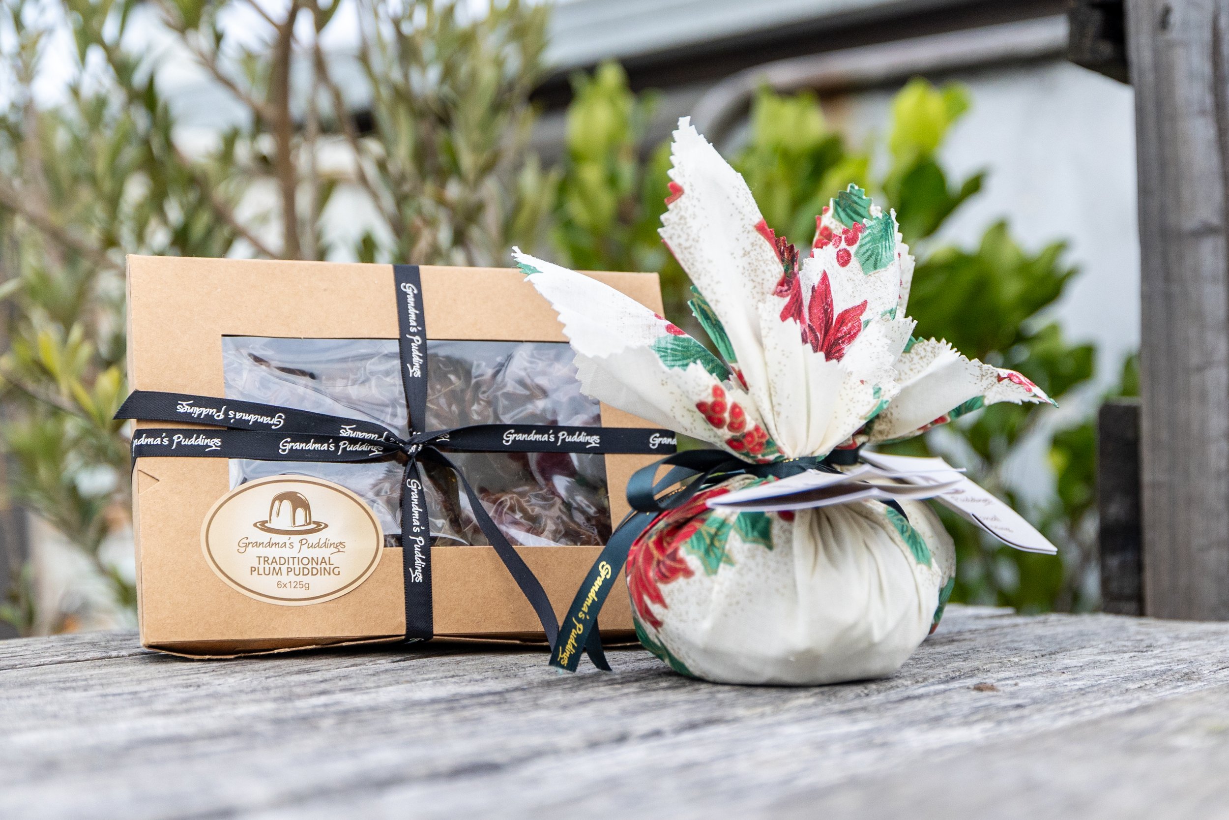 A gift box containing Grandma's Puddings Traditional Plum Pudding and a decorative festive fabric bag sitting on a wooden surface outdoors with greenery in the background.