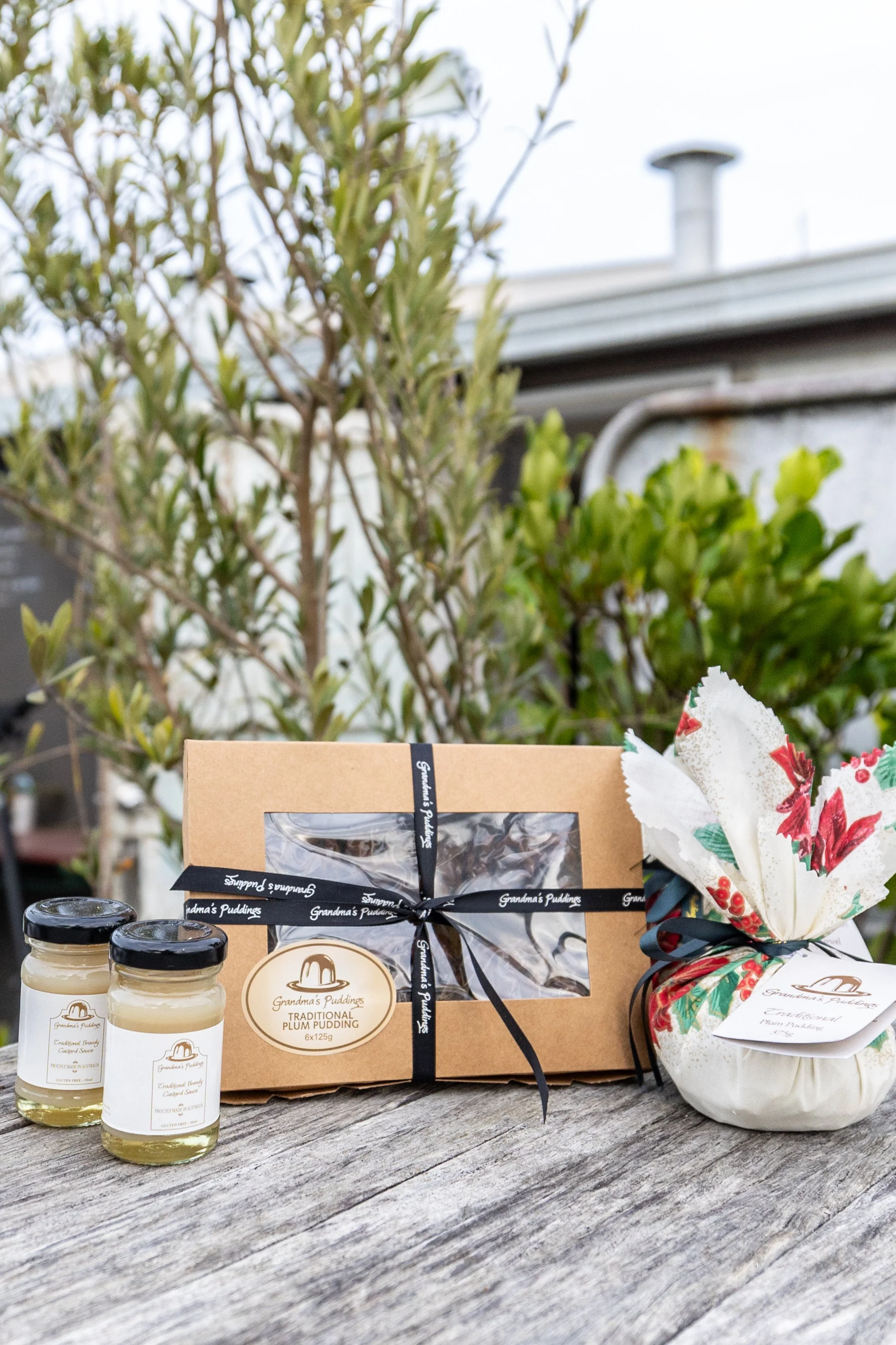 A gift box of Grandpa's Puddings traditional plum pudding with a black ribbon, two jars of caramel sauce, a floral gift bag, and some greenery in the background.