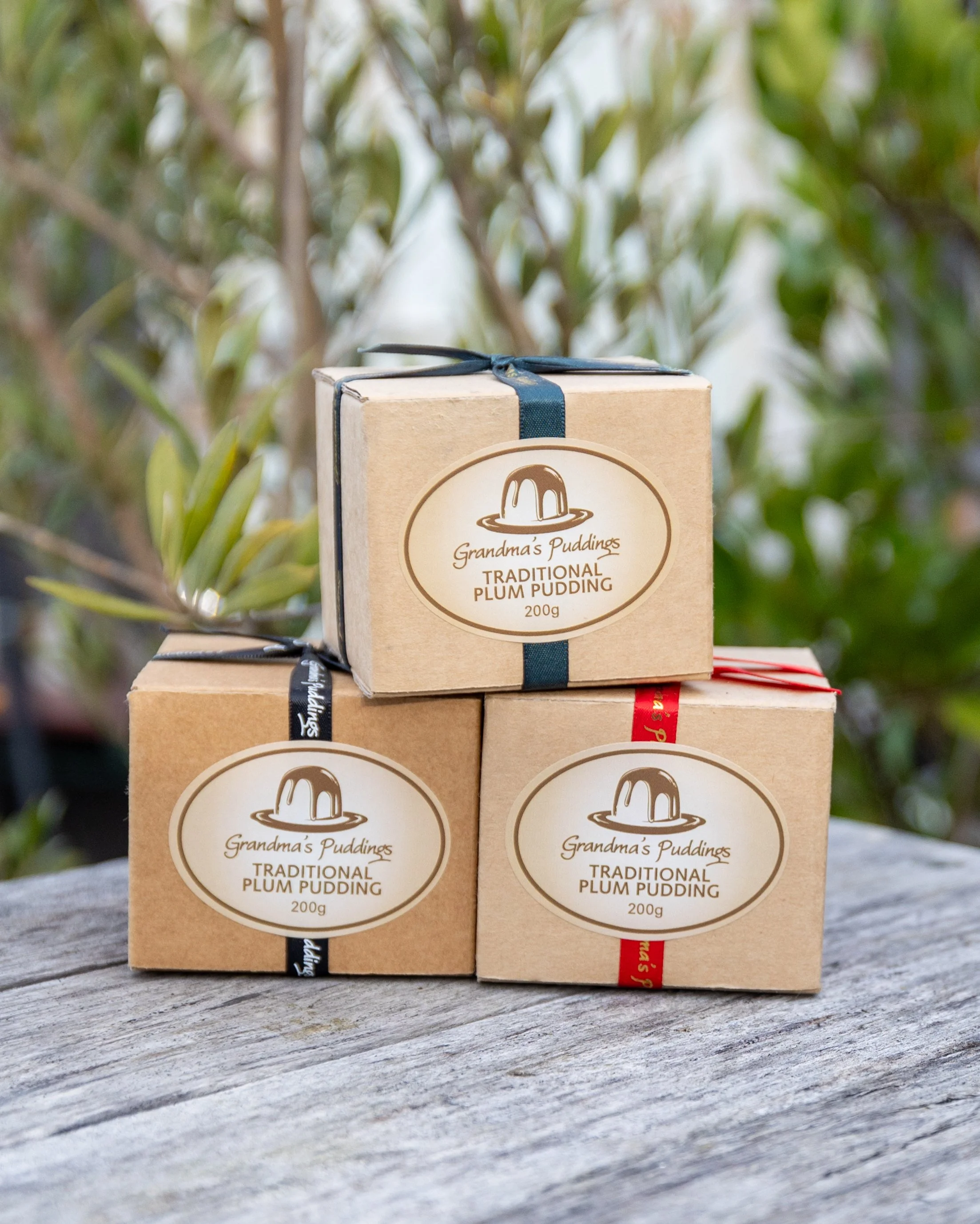 Three boxes of Grandma's Puddings Traditional Plum Pudding stacked on a weathered wooden surface with greenery in the background.