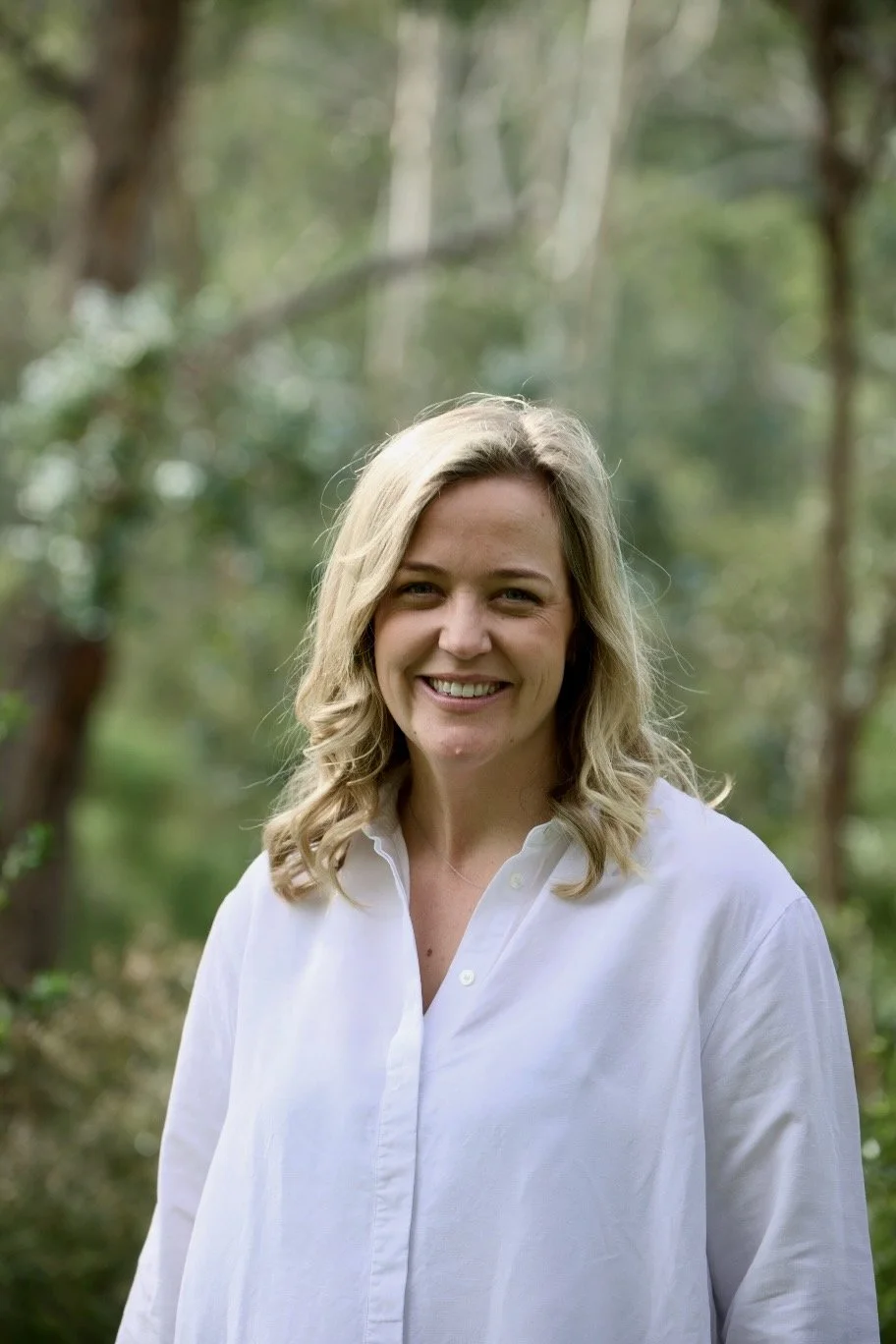 A woman with blonde curly hair smiling outdoors in a wooded area, wearing a white button-up shirt.