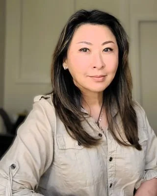 A woman with shoulder-length dark hair wearing a beige button-up shirt, standing indoors with a neutral background.