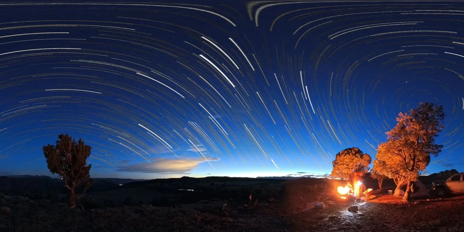 Star trails in the night sky over a campsite with trees, a fire, and tents.