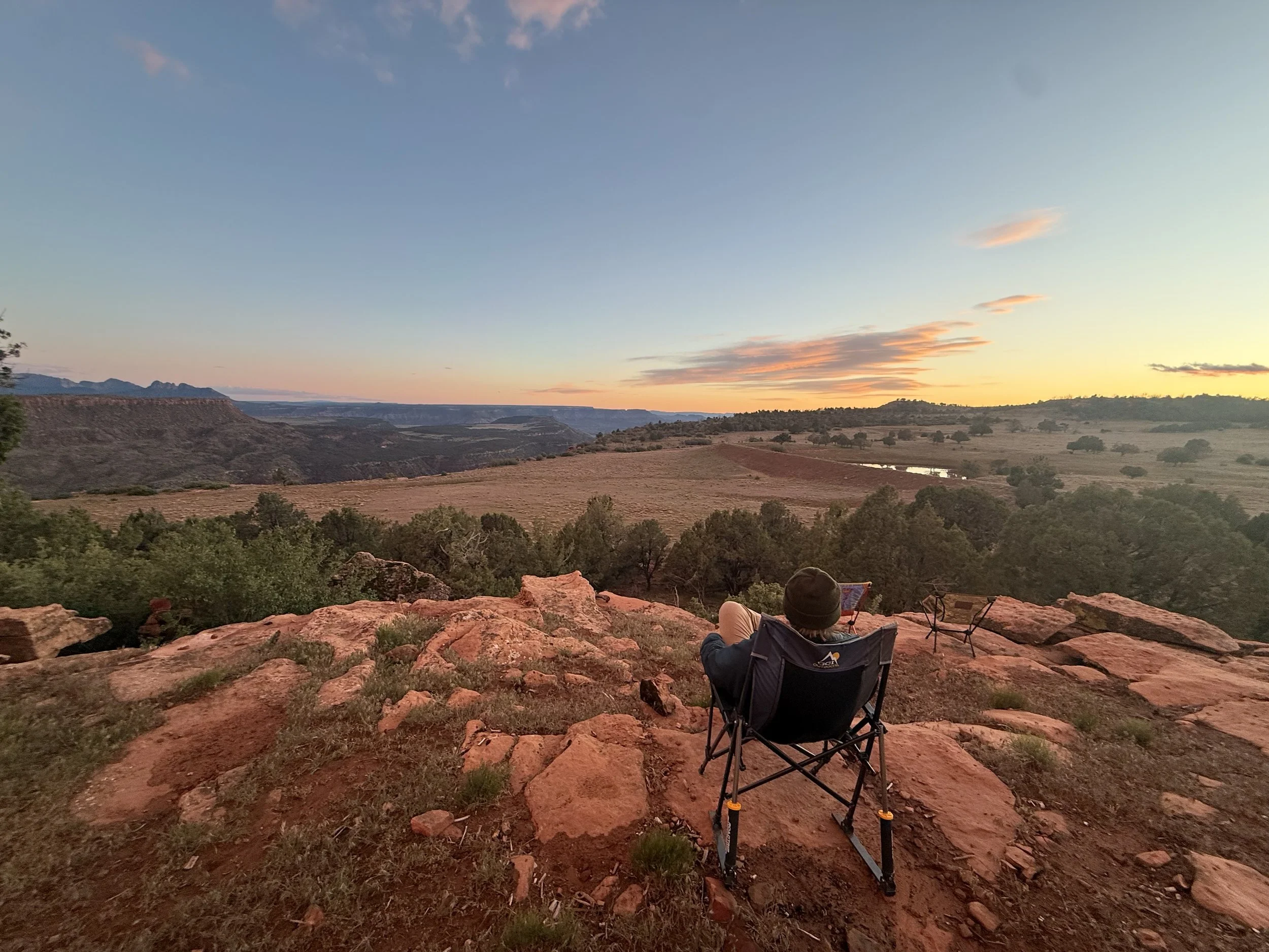 A person sitting in a camping chair on red rock, overlooking a scenic landscape during sunset, with trees, a river, and distant hills.