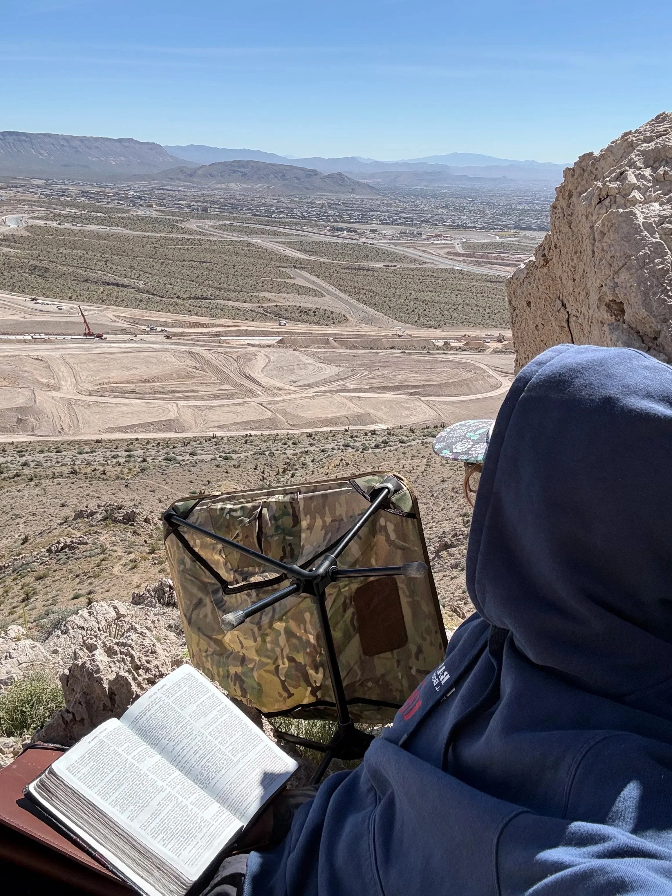 A person sitting on a rock ledge overlooking a desert landscape with mountains in the distance. The person is reading a large book, with a camouflage-patterned bag and an open book on a small table nearby, and a wide view of the valley and winding roads below.