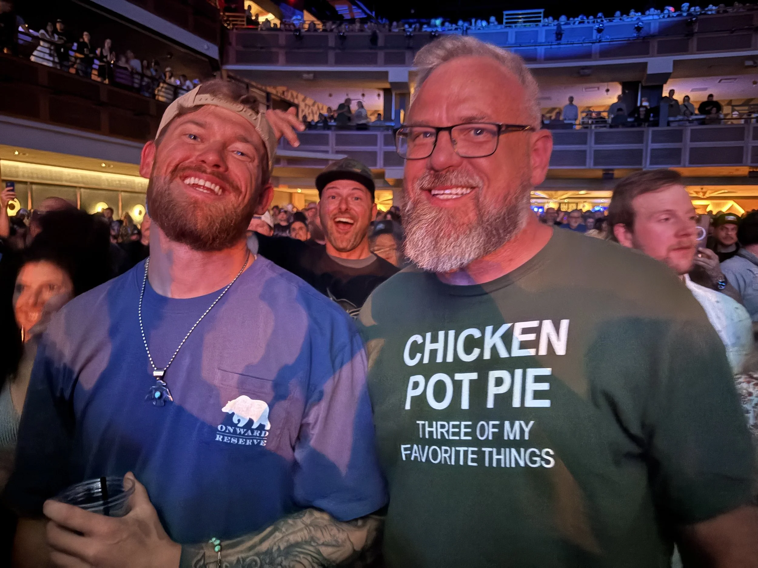 Two men smiling at a crowded indoor event, with multiple levels and blue lighting in the background. One man has a beard, glasses, and a gray shirt with text; the other man has a beard, wearing a blue shirt with a logo, and a headband.