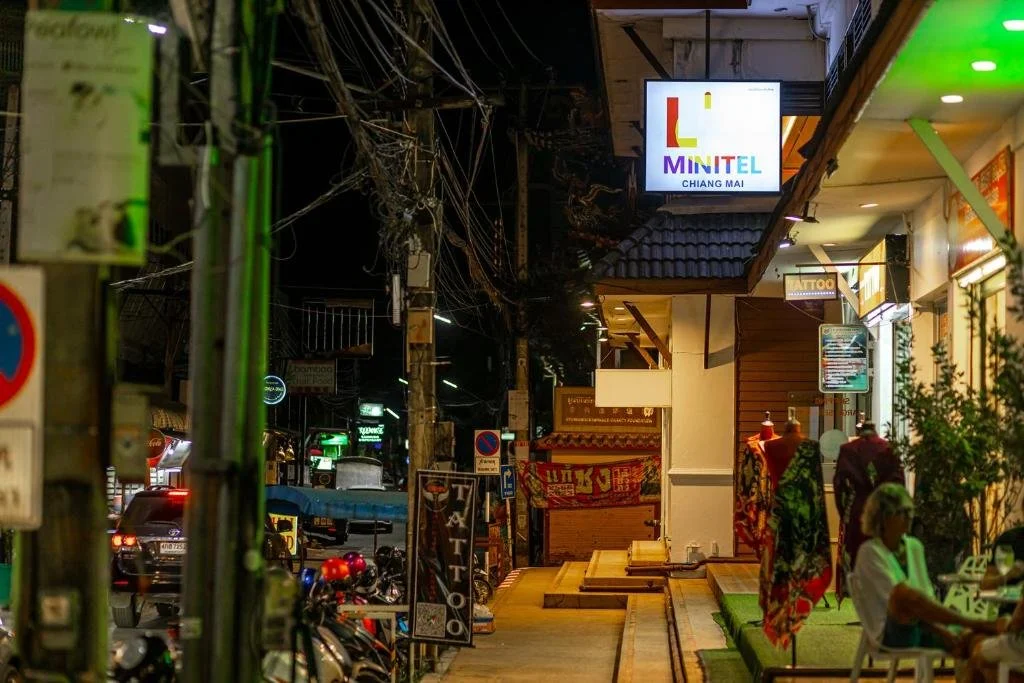 Nighttime street scene in Chiang Mai, Thailand, with signs including Minitel and tattoo shops, and mannequins in colorful clothing outside shops.
