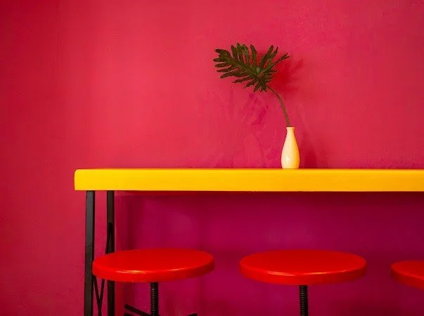 A red wall with a yellow counter or table, three red bar stools, and a white vase with a green plant.