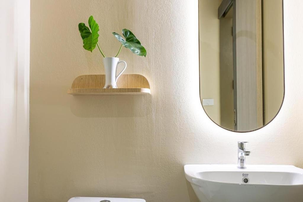 A bathroom interior with a white sink, an oval mirror, a wall-mounted wooden shelf holding a white pitcher with green leaves, and a beige wall.