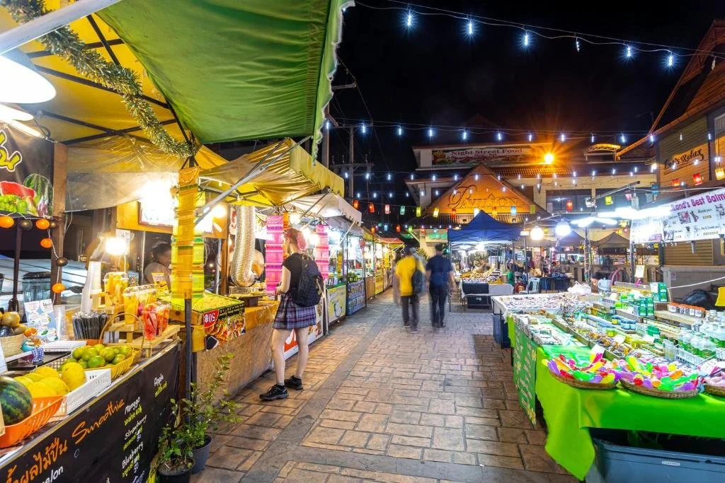 Night market with food stalls on both sides, decorated with string lights, and a few people shopping.