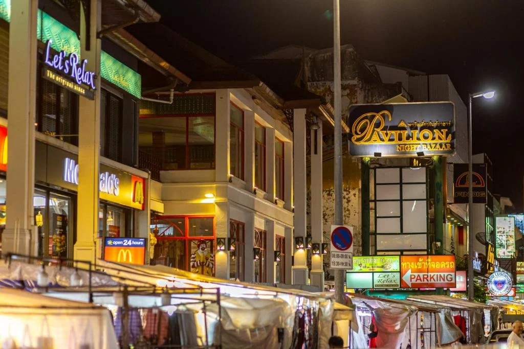 Night scene of a street with illuminated storefronts and signs, including a McDonald's, a sign for Pavilion Night Bazaar, and various other businesses. Street stalls with tents are visible at the bottom.