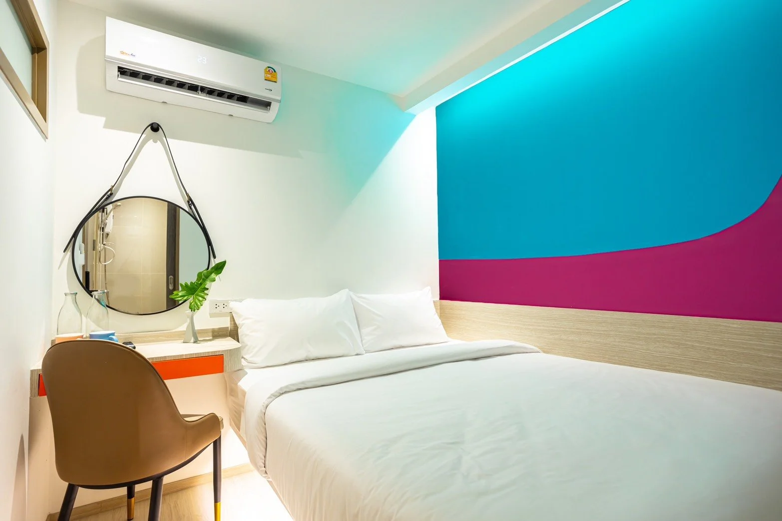 Hotel room with white bed, colorful blue and pink accent wall, round mirror, wooden desk with a chair, and air conditioning unit.