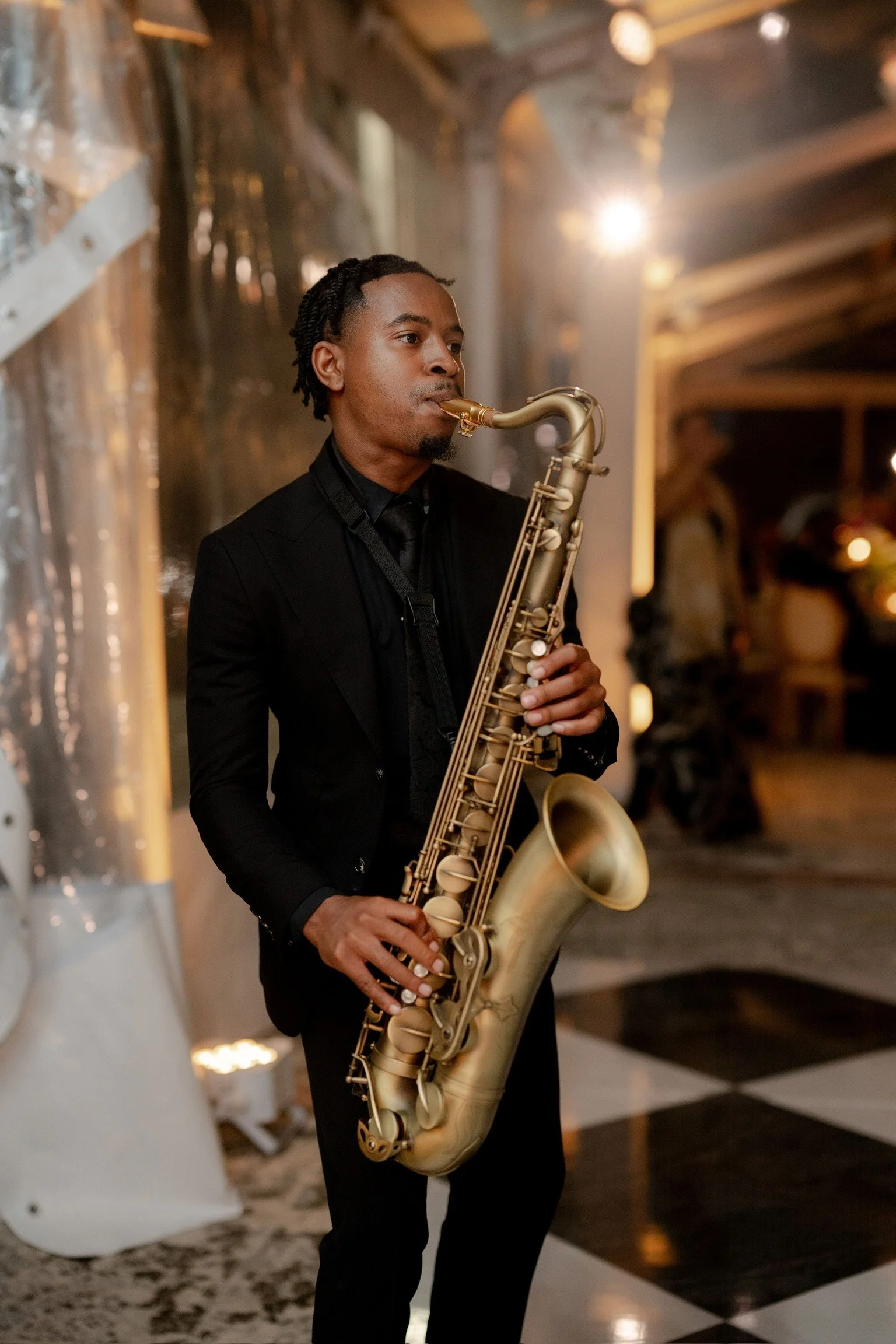 A man dressed in a black suit playing a gold saxophone indoors with warm lighting and modern decor.