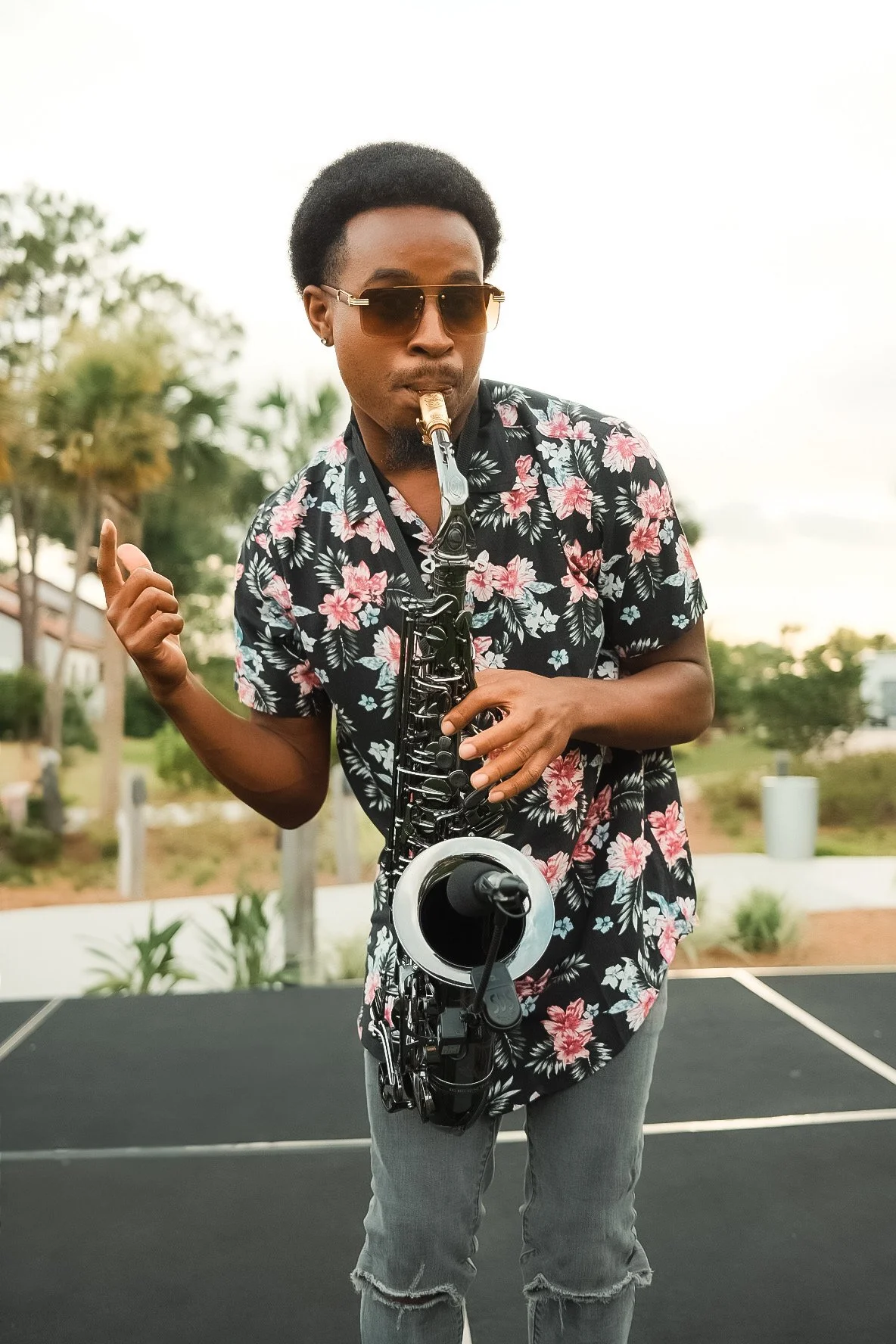 A man wearing sunglasses and a floral shirt is playing a saxophone outdoors, with trees and a white sky in the background.