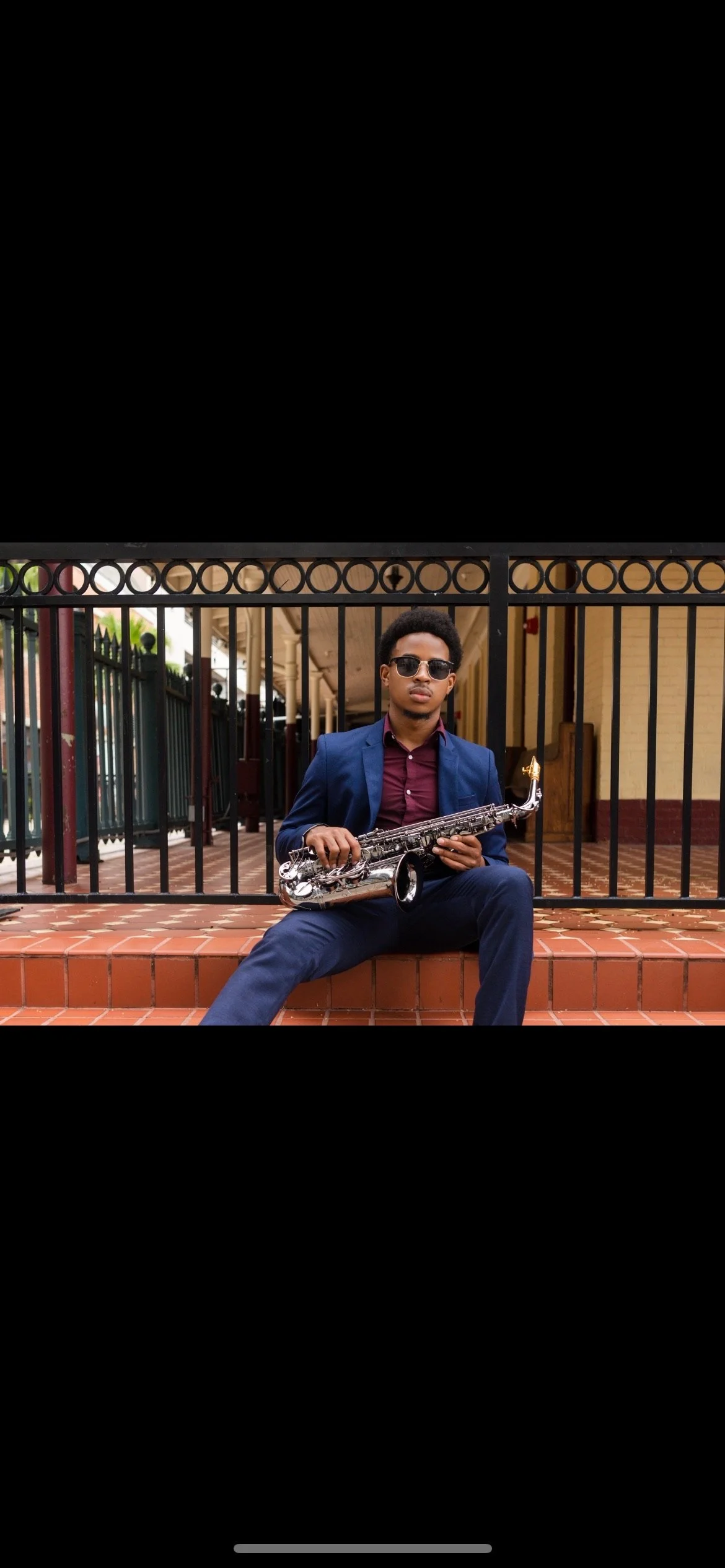 Young man sitting on brick steps, holding a saxophone, wearing sunglasses, a blue suit, and a maroon shirt, outdoors near a yellow building with black metal fence.