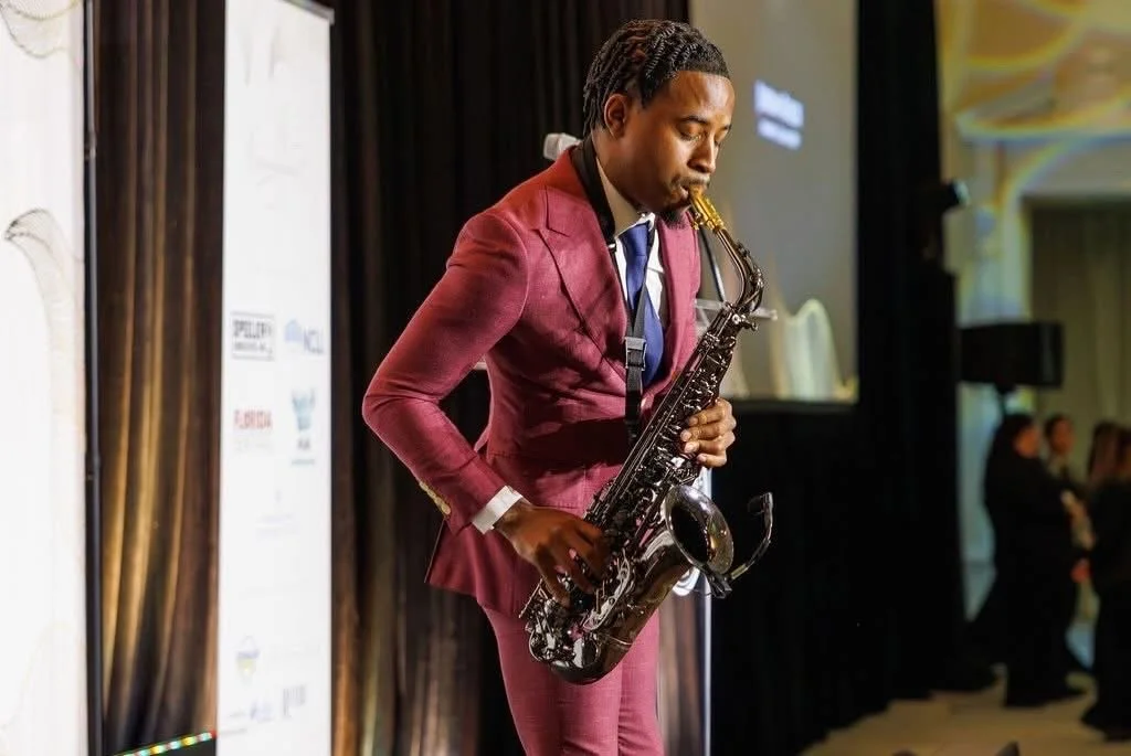 A man wearing a burgundy suit plays a saxophone on stage during an event.
