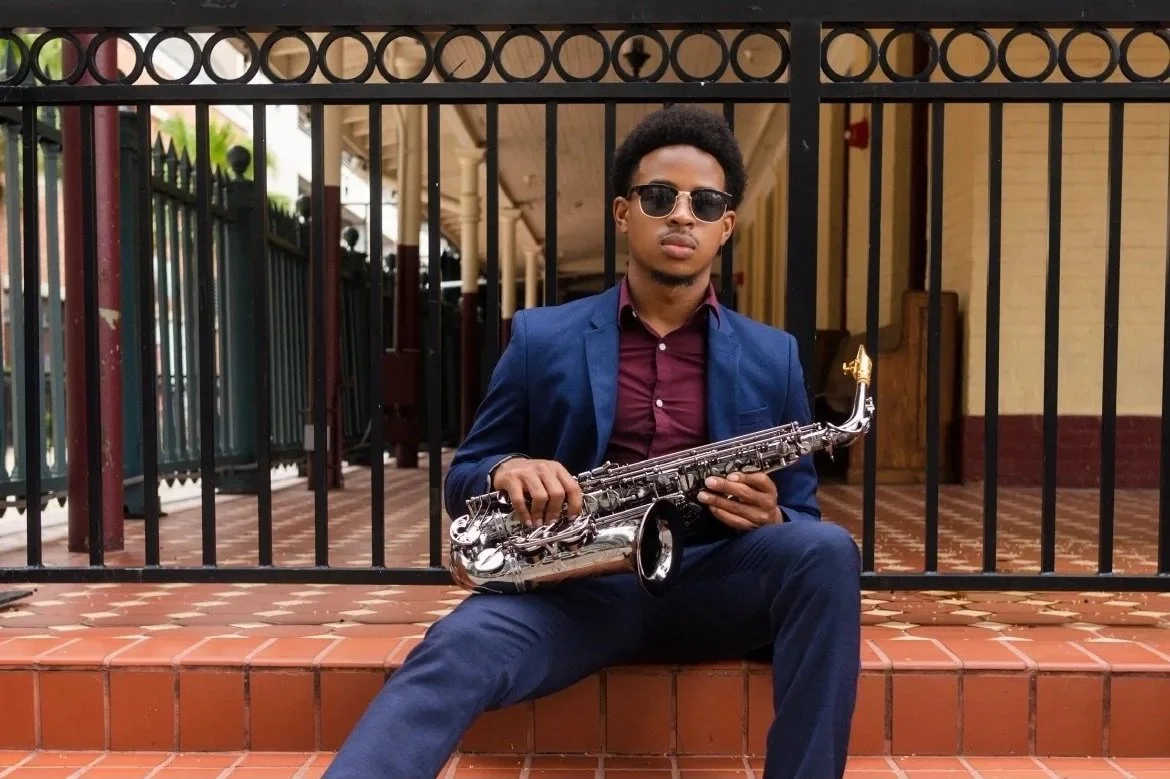 A young man in a blue suit and sunglasses sitting on brick stairs holding a silver saxophone.