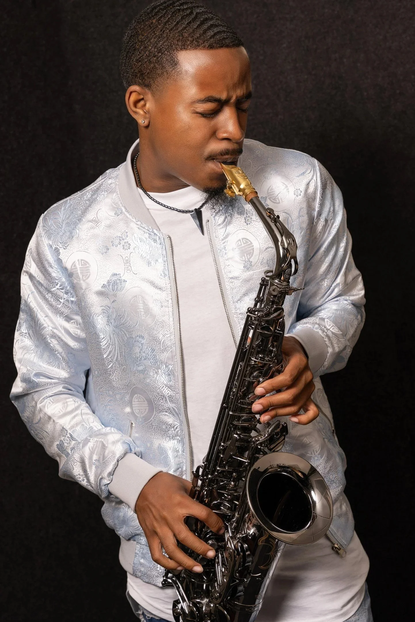 A young man playing a saxophone against a black background, wearing a white satin jacket with intricate patterns.