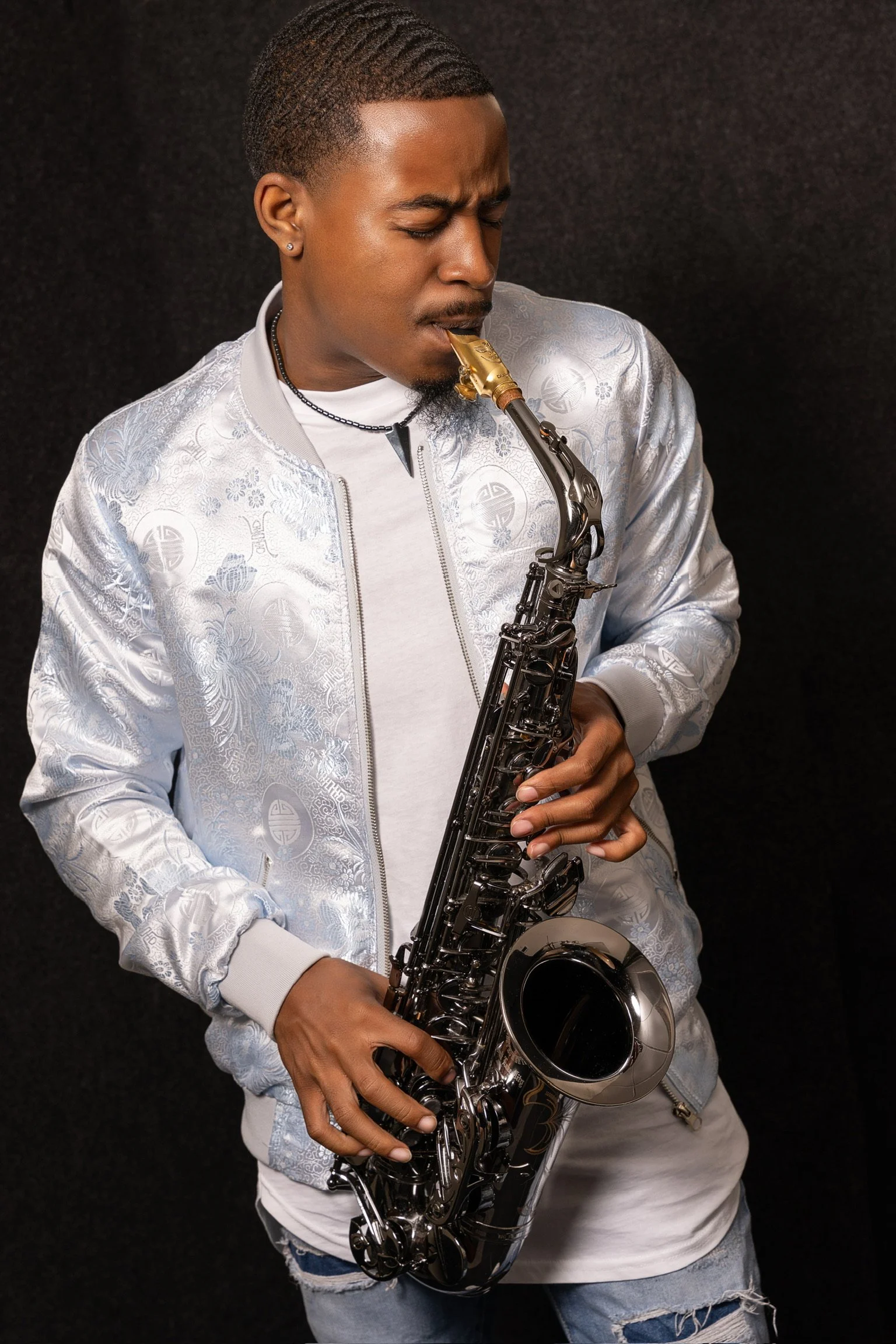 A young man with short curly hair plays a black and gold saxophone against a black background. He is wearing a white patterned jacket over a plain white t-shirt, with ripped jeans, and has earrings and necklaces.