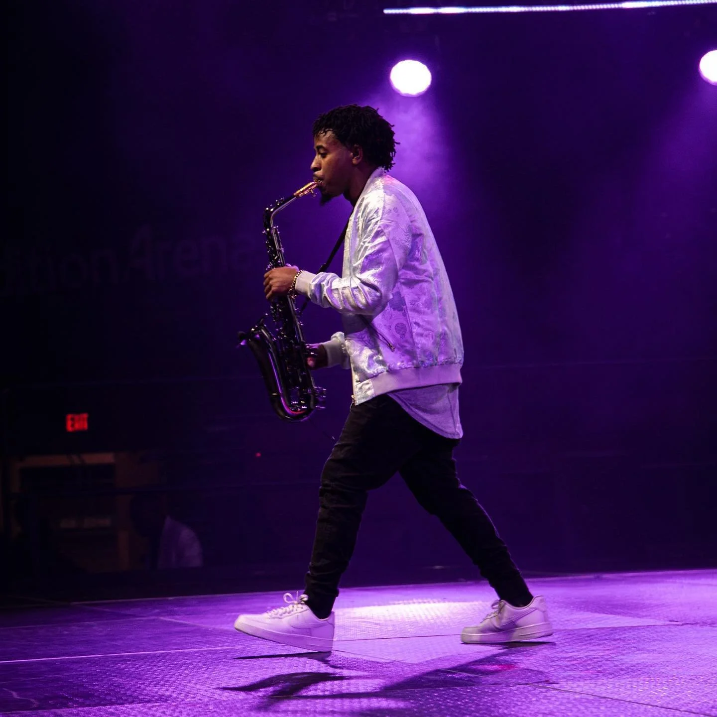 A young man playing a saxophone on a stage illuminated by purple lights, wearing a silver jacket, black pants, and white sneakers.